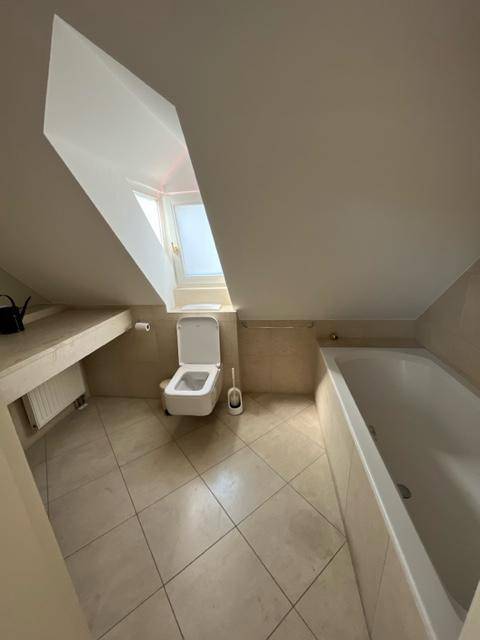 Modernes Badezimmer mit beigen Fliesen, wandhängendem WC und einer Badewanne unter einem Dachfenster.