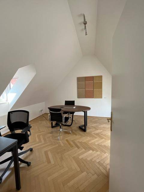 Helles Büro mit schrägen Decken, Fischgrätparkett und einem runden Holztisch mit Bürostühlen.