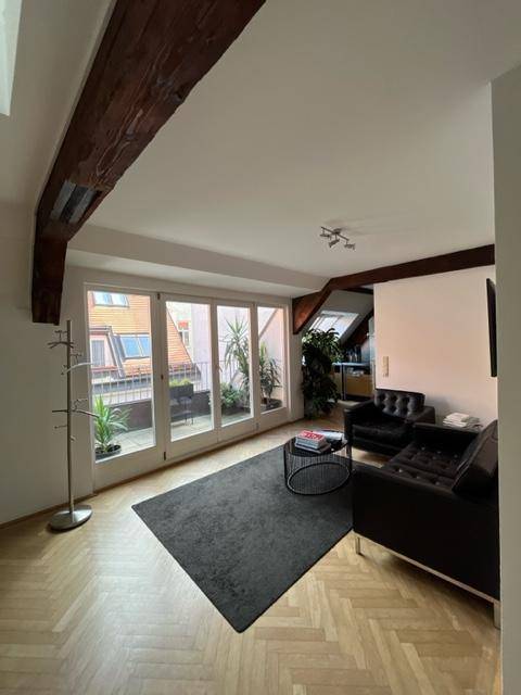 Helles Wohnzimmer mit schwarzen Ledersofas, Teppich und großen Glastüren, die auf einen Balkon führen.