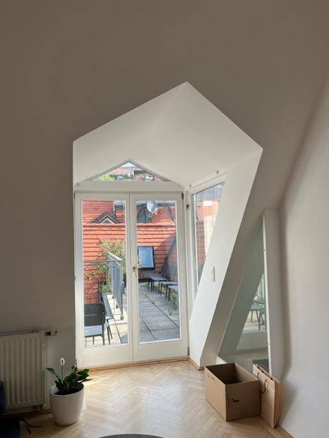 Innenansicht eines Raumes mit schrägen Wänden und großen Fenstern, die auf eine Dachterrasse führen.