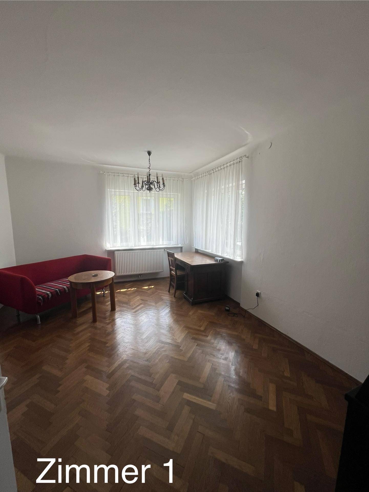 Helles Wohnzimmer mit Fischgrätparkett, rotem Sofa und einem Schreibtisch am Fenster.