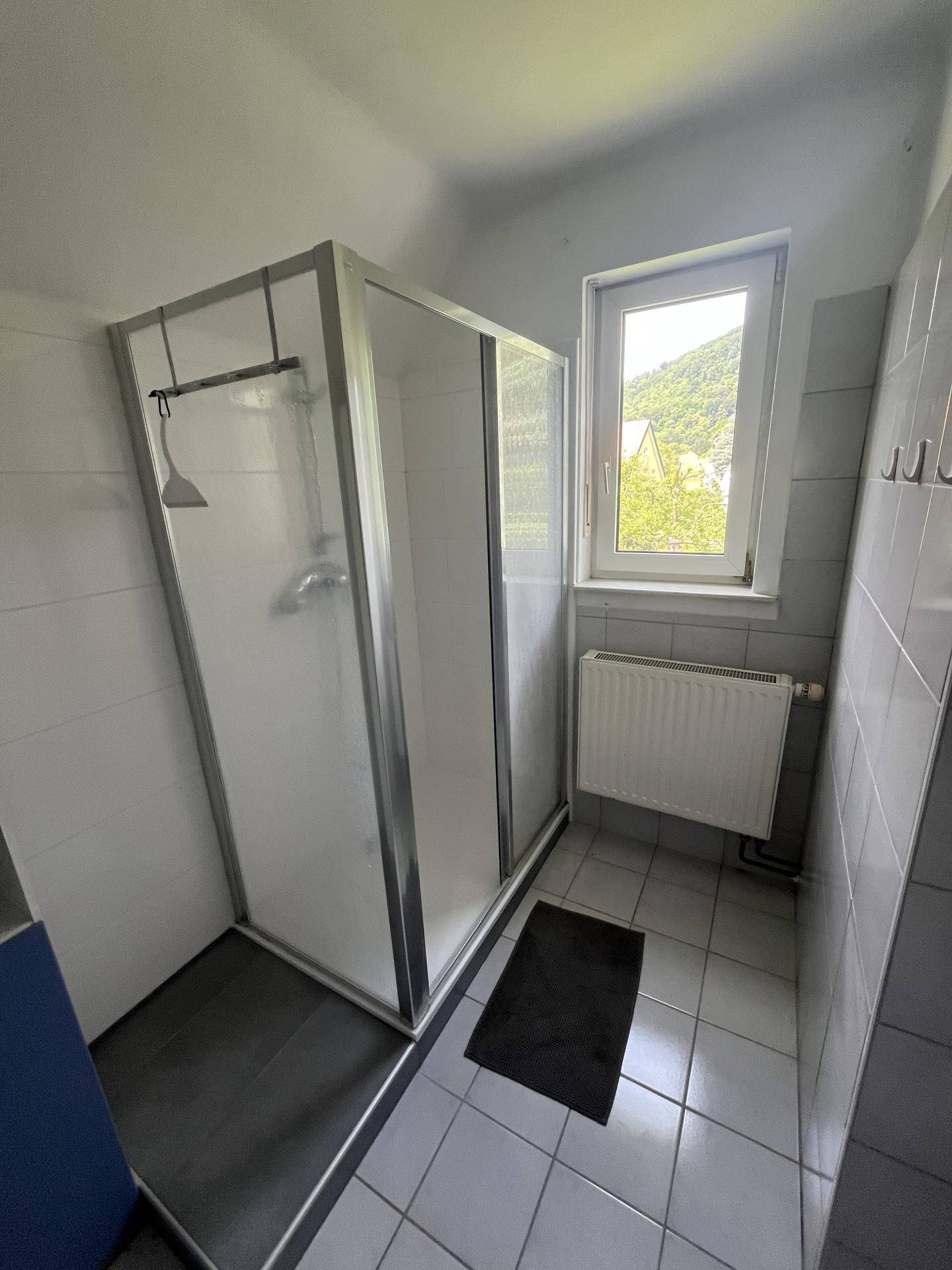 Badezimmer mit Duschkabine, Fliesenboden und Fenster, das viel Tageslicht hereinlässt.