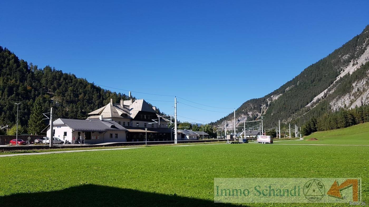 Ein Bahnhofsgebäude mit Gleisen im Vordergrund, umgeben von einer weiten grünen Wiese und Bergen.