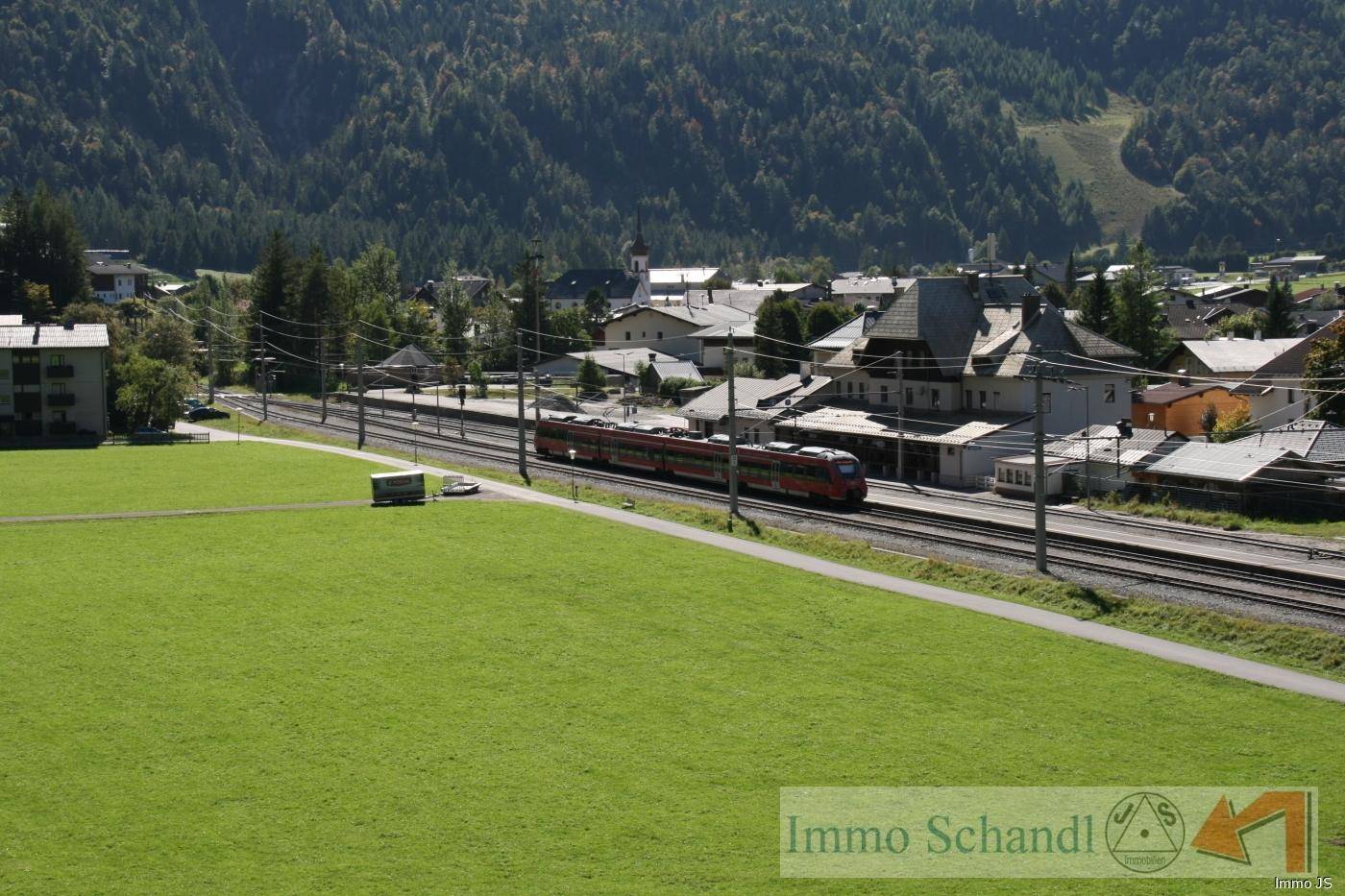 Eine erhöhte Ansicht von Bahngleisen mit einem Zug, einer grünen Wiese und einem Dorf vor Bergkulisse.