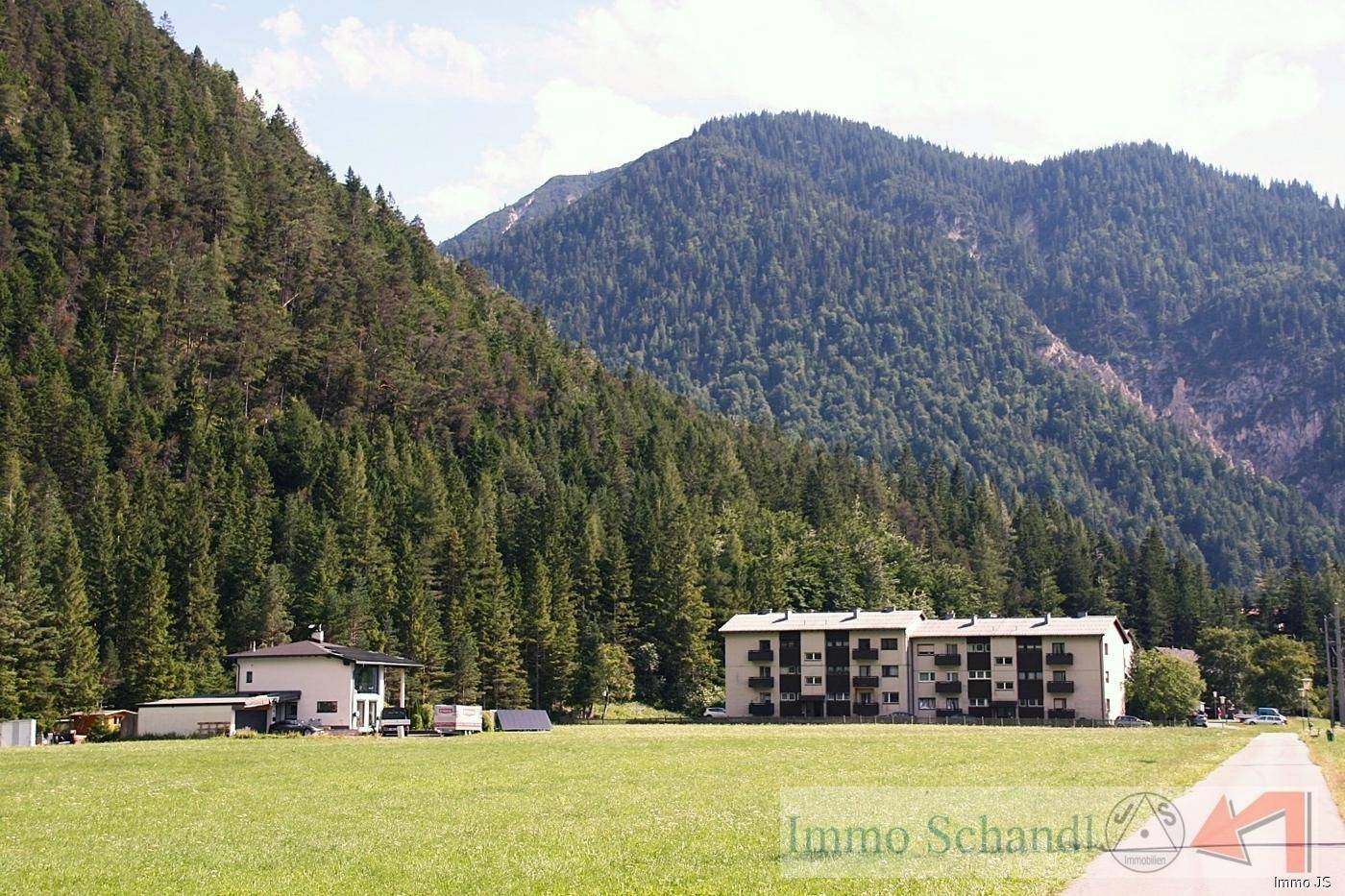 Zwei Gebäude, ein Haus und ein Mehrfamilienhaus, eingebettet in eine grüne Landschaft vor bewaldeten Bergen.