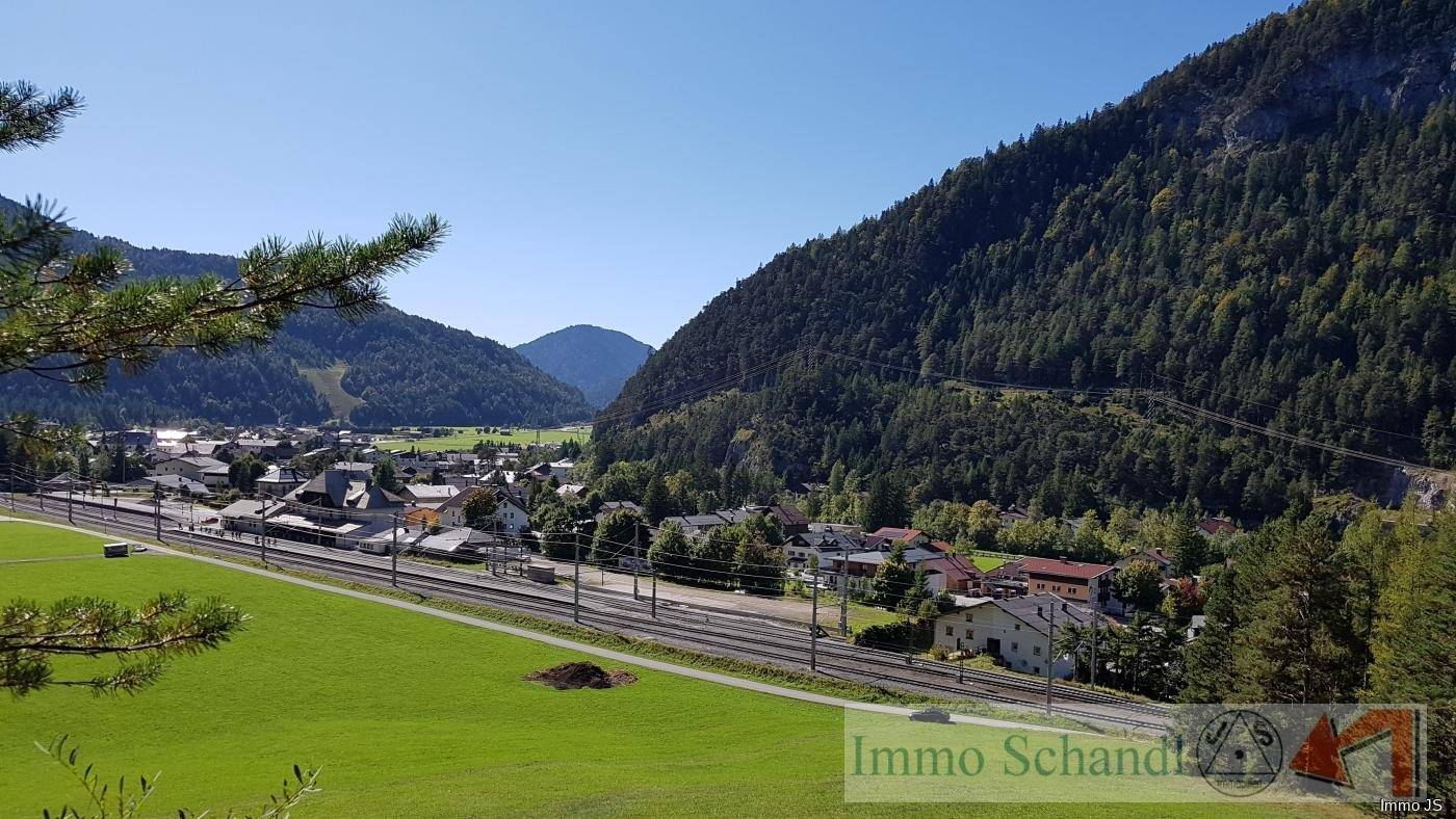 Panoramablick auf ein Dorf in einem Tal, umgeben von bewaldeten Bergen und Bahngleisen im Vordergrund.