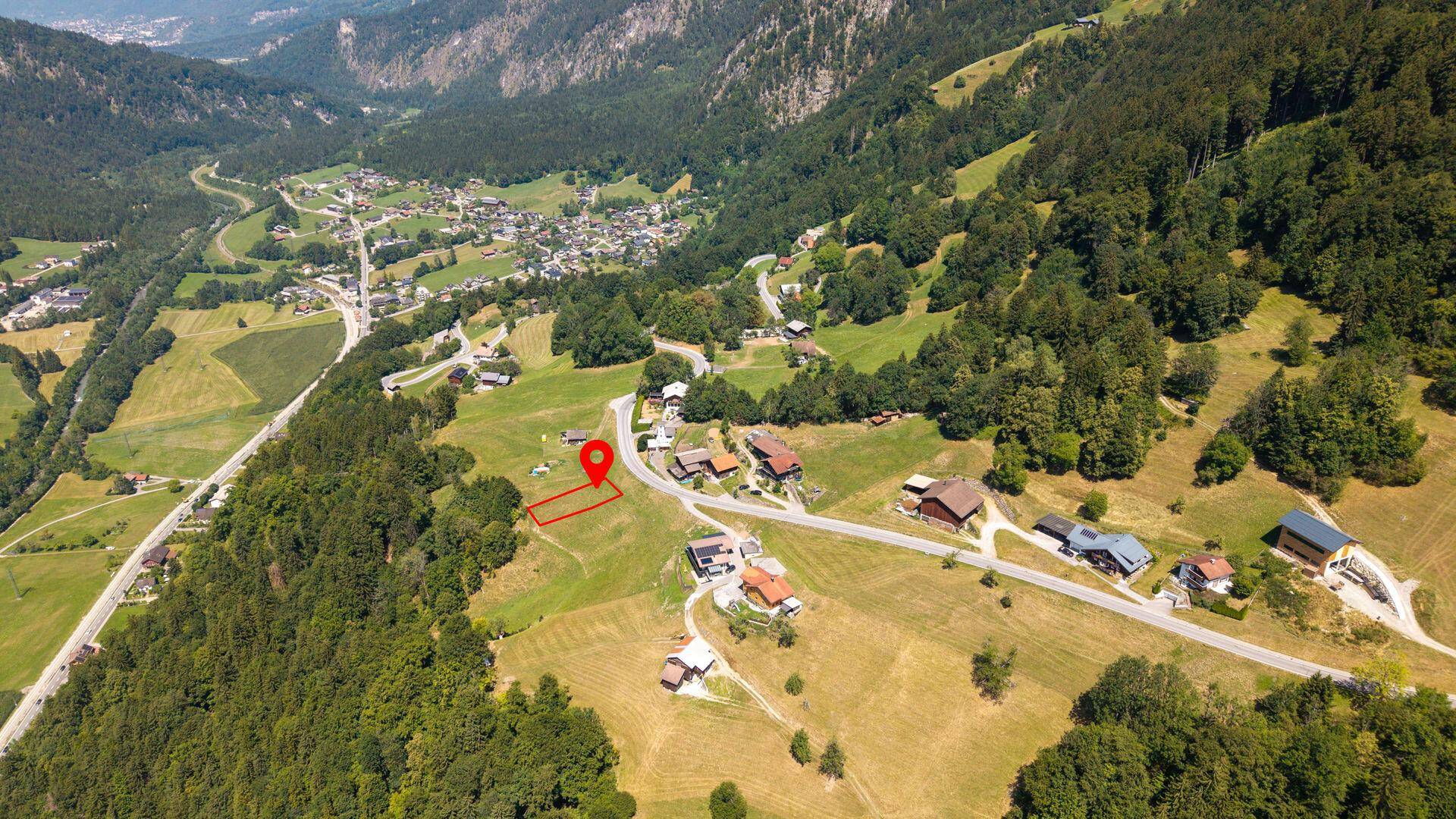 Weitläufige Luftaufnahme eines Bergdorfes mit rot markiertem Grundstück in idyllischer Umgebung.