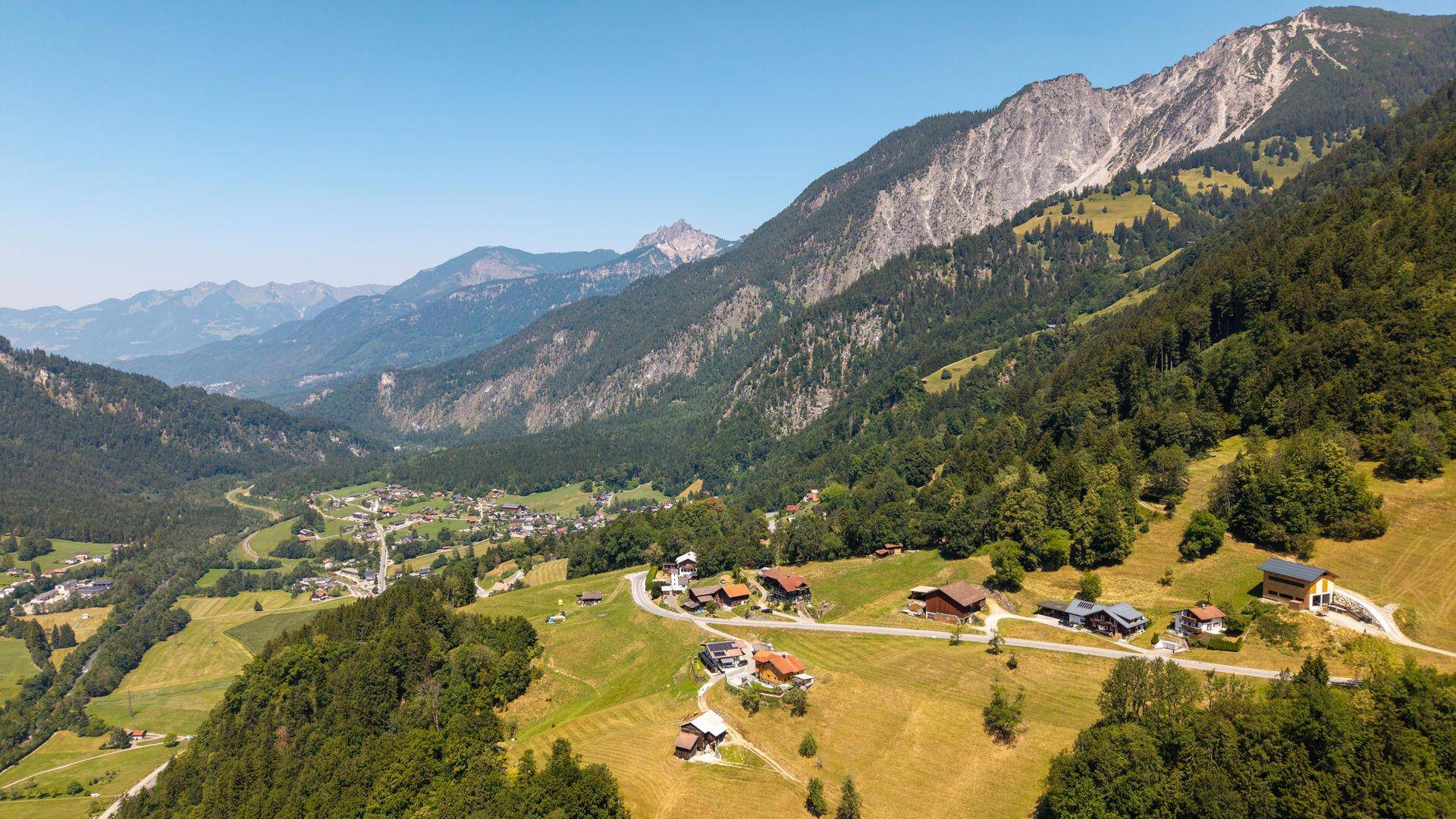 Malerische Luftaufnahme eines Dorfes in einem weiten Tal, umgeben von majestätischen Bergen unter blauem Himmel.