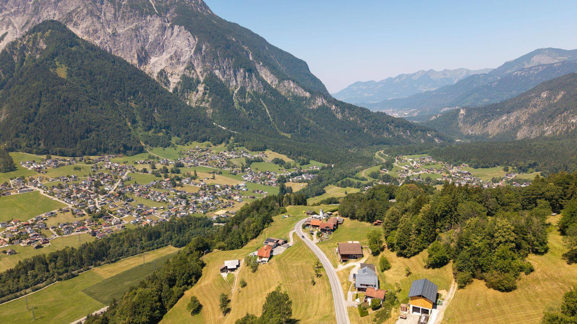 Panoramablick auf ein idyllisches Dorf in einem Bergtal, umgeben von hohen Gipfeln und grünen Wäldern.
