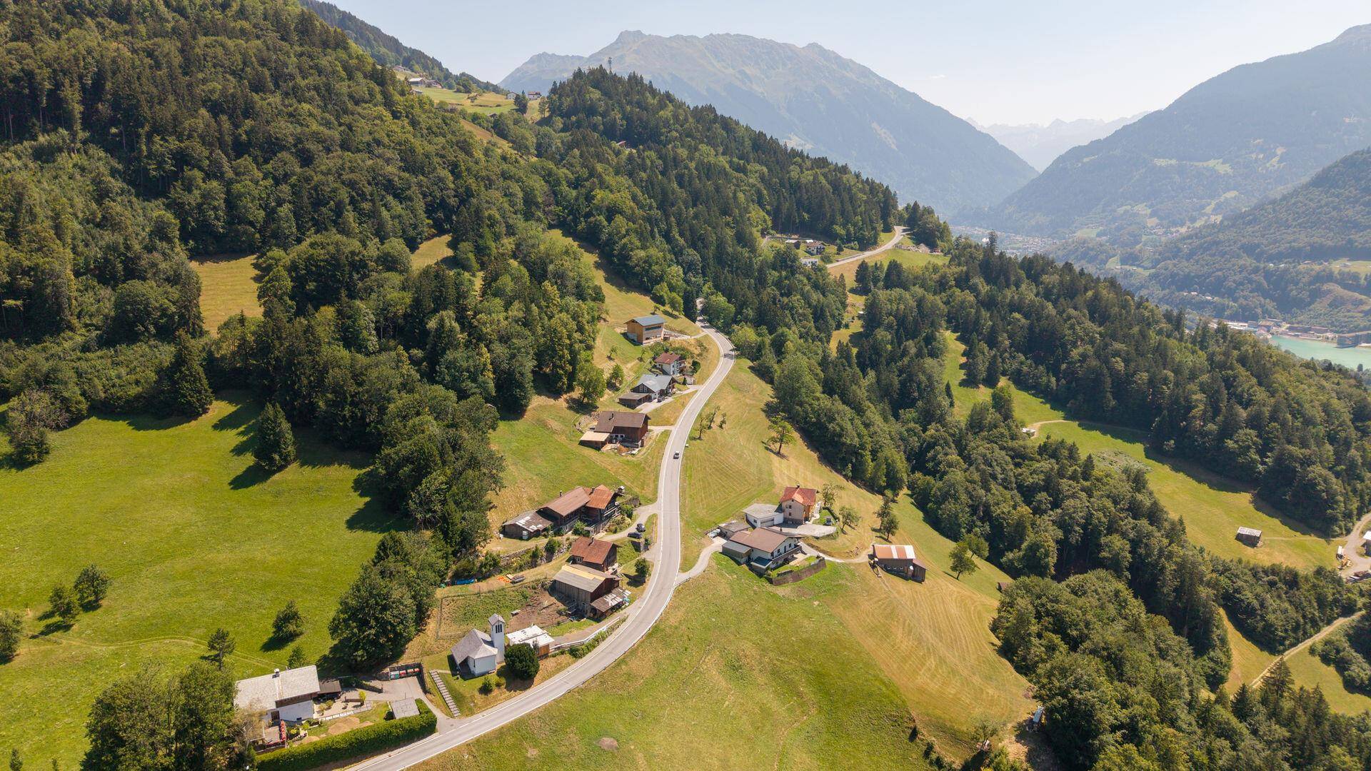Luftaufnahme eines Bergdorfes mit einer sich schlängelnden Straße, eingebettet in eine grüne Hügellandschaft.