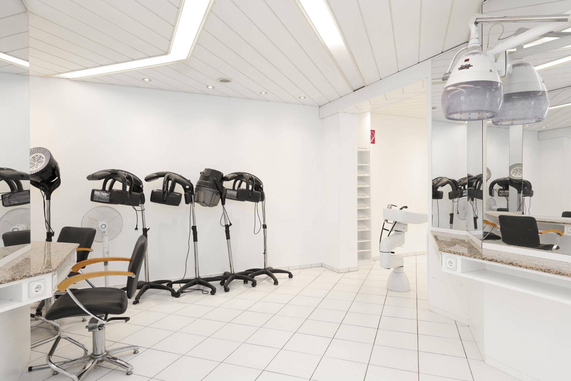 Friseursalon mit mehreren Trockenhauben, Spiegeln und Arbeitsplätzen auf weiß gefliestem Boden.
