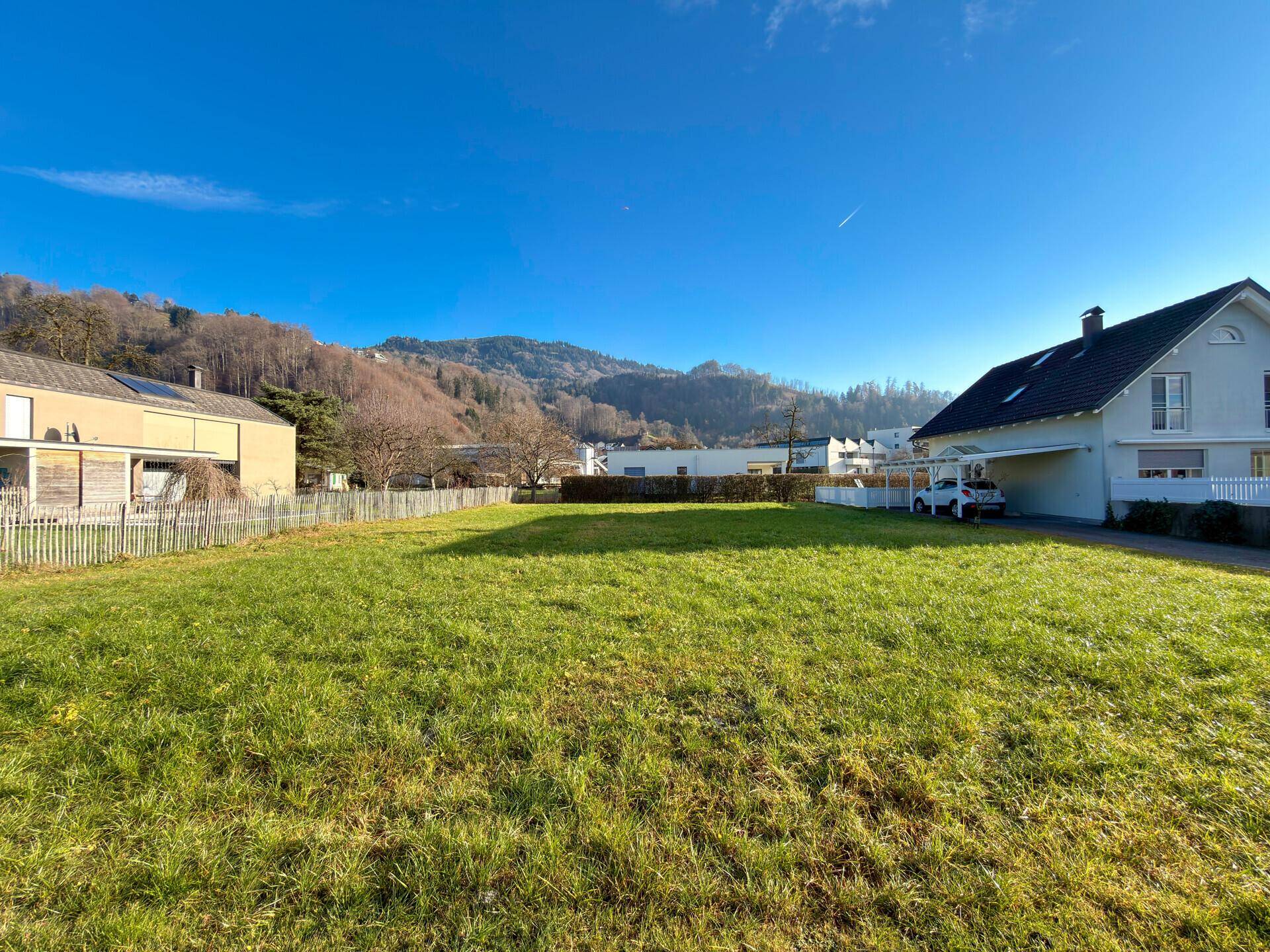 Sonniger Blick über eine grüne Wiese mit angrenzenden Häusern und bewaldeten Bergen im Hintergrund.