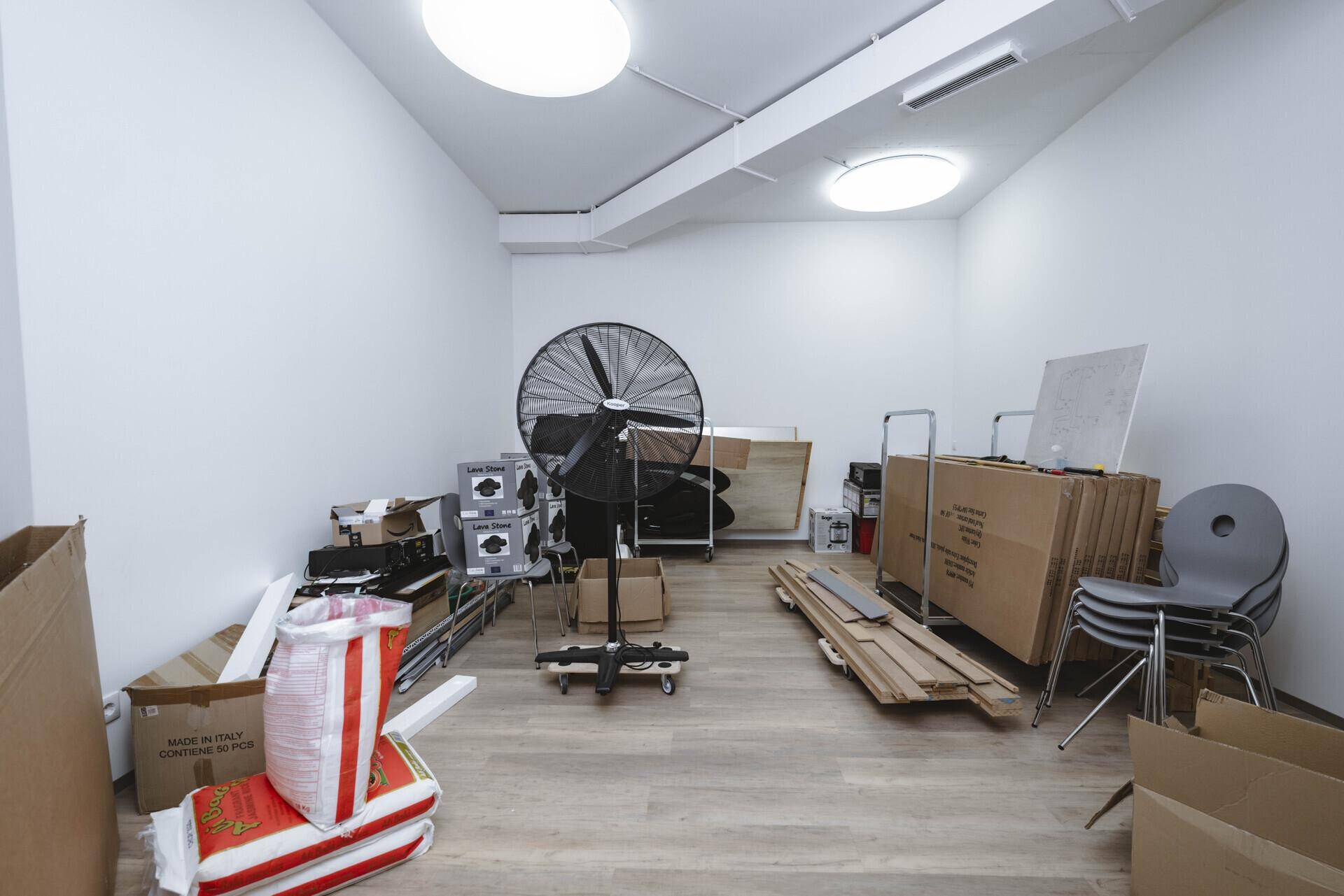 Leerer Lagerraum mit Kartons, einem großen Ventilator und hellem Laminatboden.