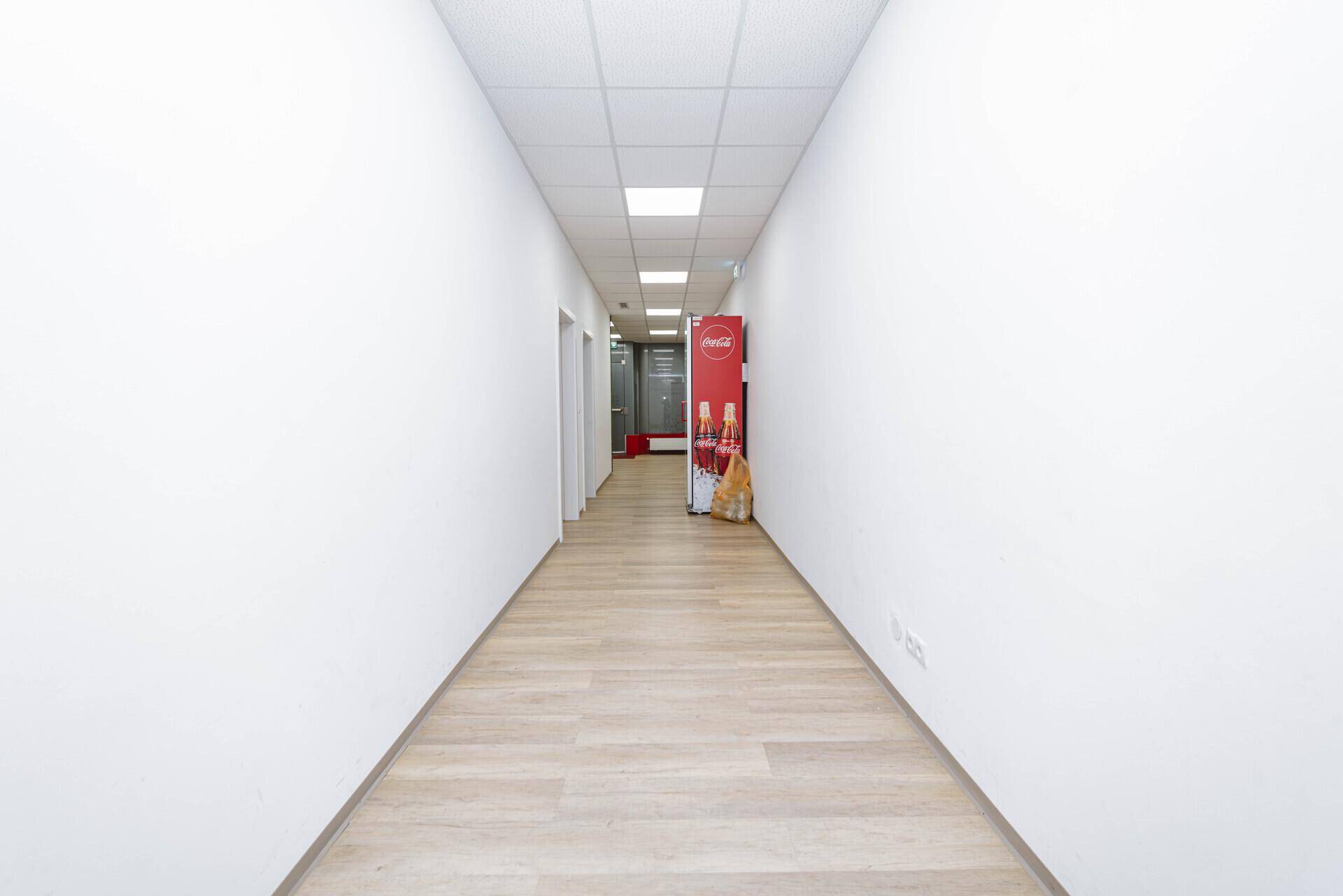 Langer, heller Flur mit weißen Wänden, Laminatboden und einem Getränkekühlschrank am Ende.