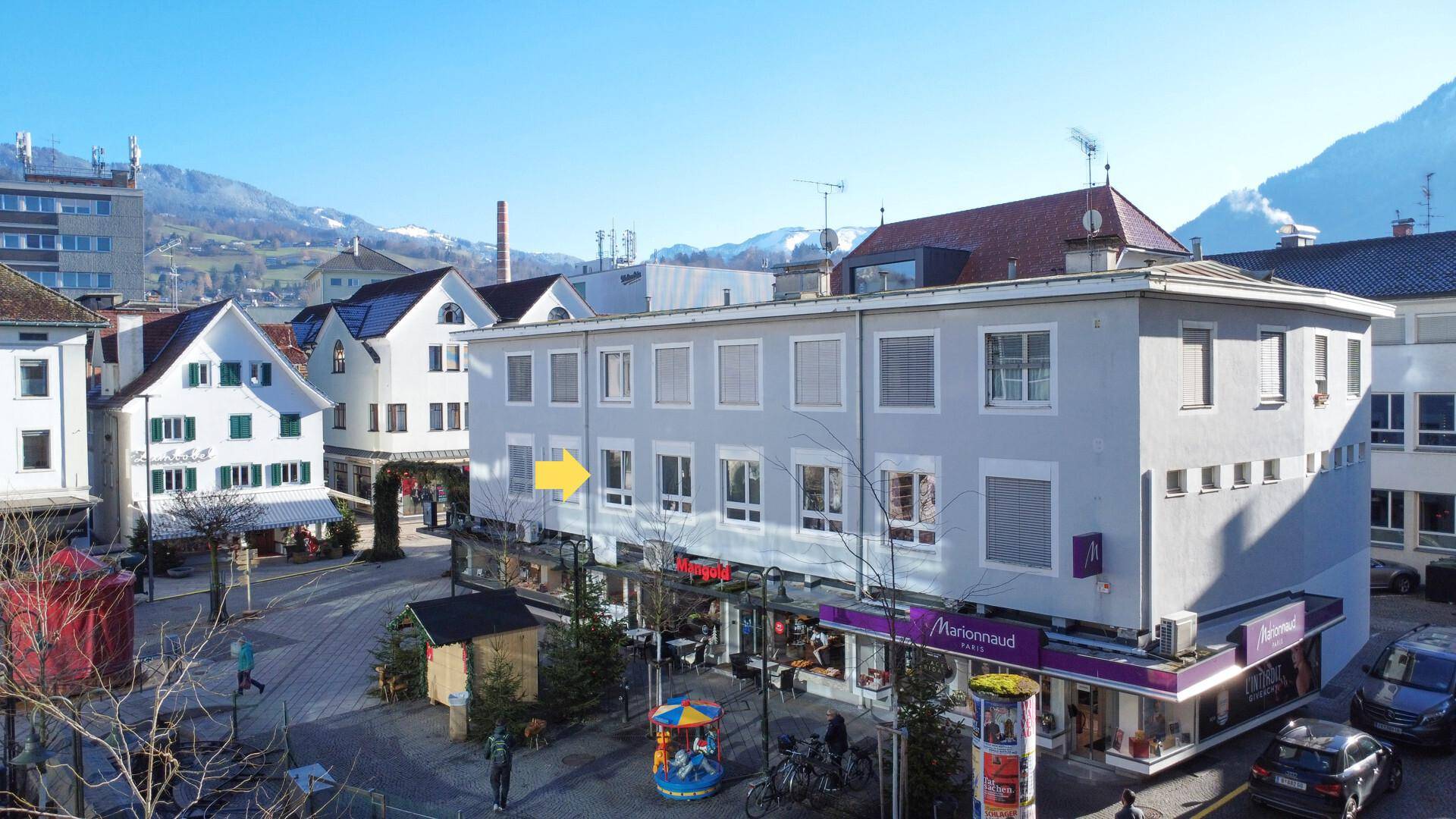 Gebäudeansicht in einer belebten Stadtumgebung mit Geschäften im Erdgeschoss und Bergen im Hintergrund.