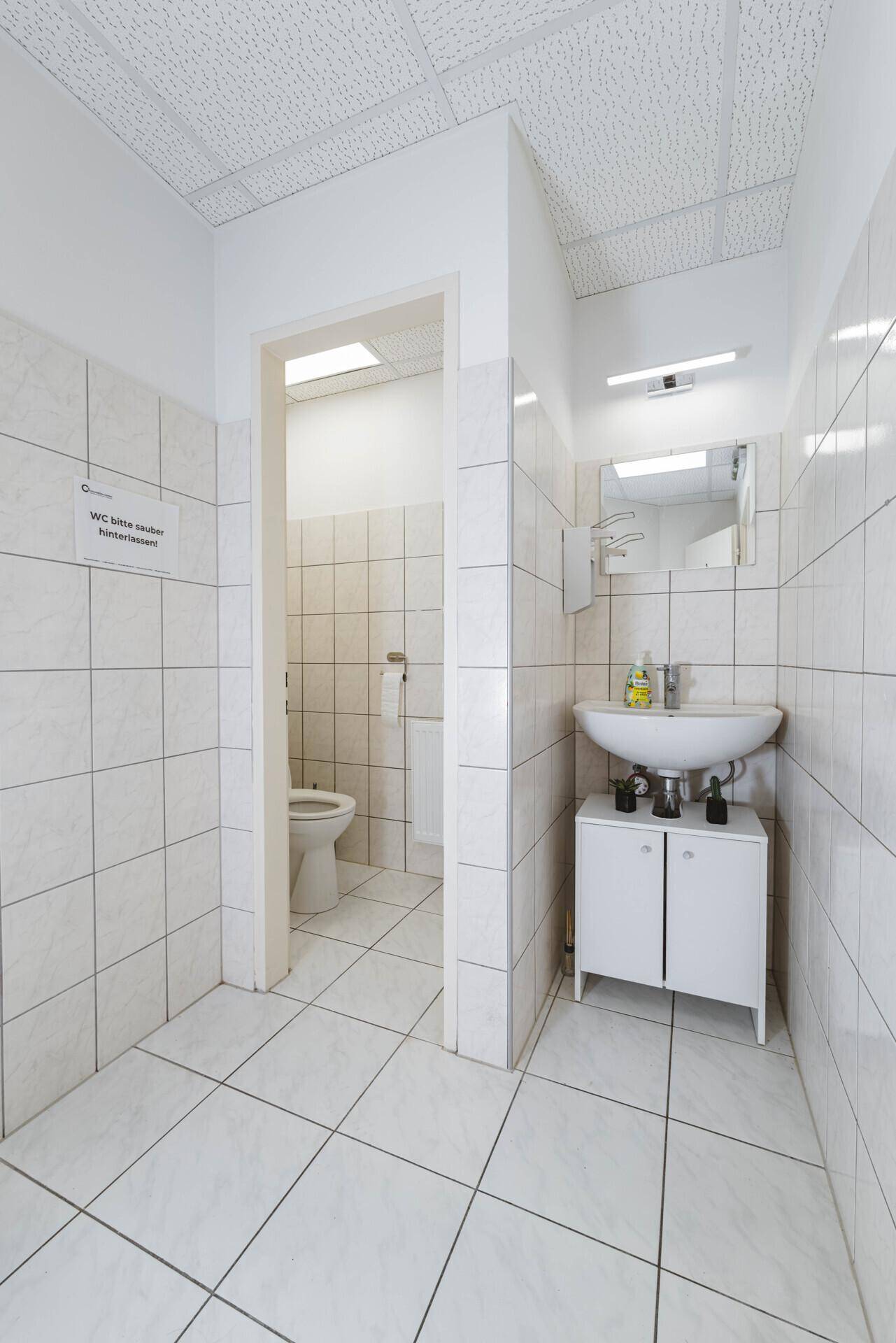 Gefliestes WC mit Waschbecken, kleinem Unterschrank und Toilette im Hintergrund.