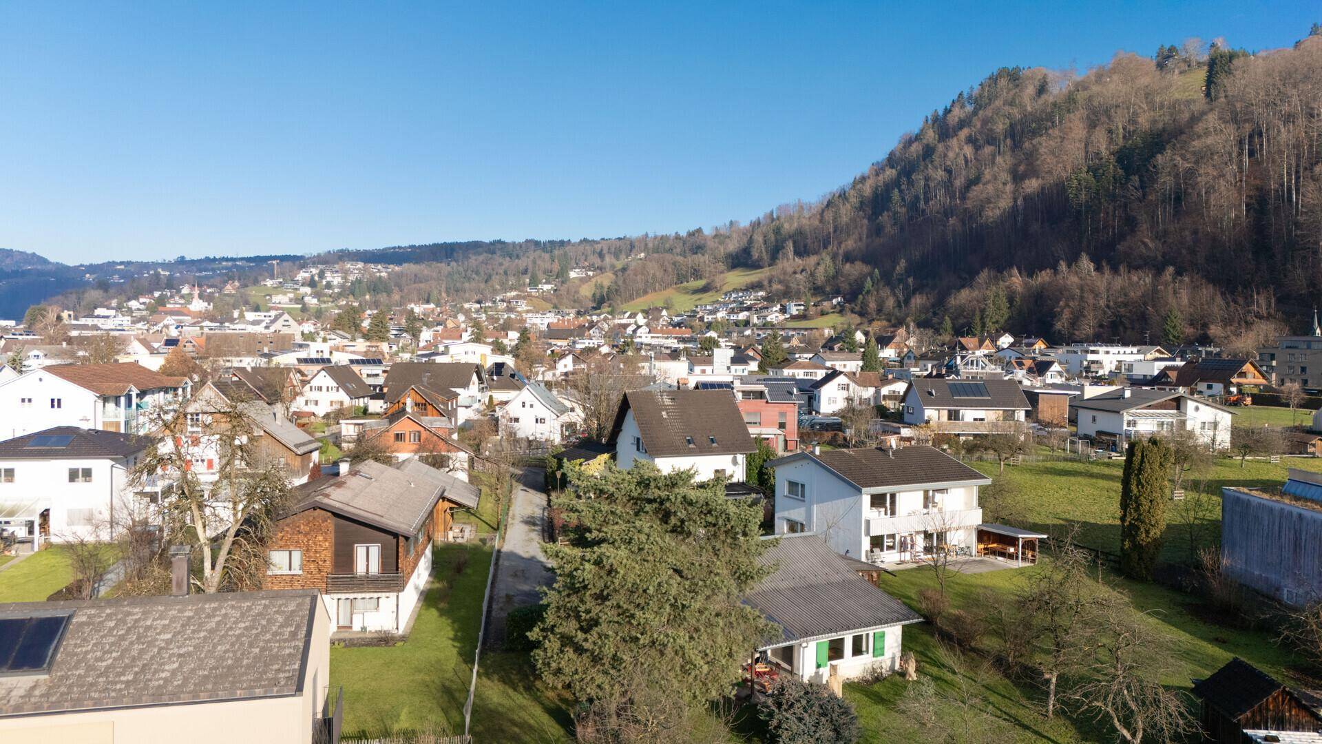 Panoramablick aus der Vogelperspektive auf eine Wohnsiedlung mit bewaldeten Hügeln im Hintergrund.