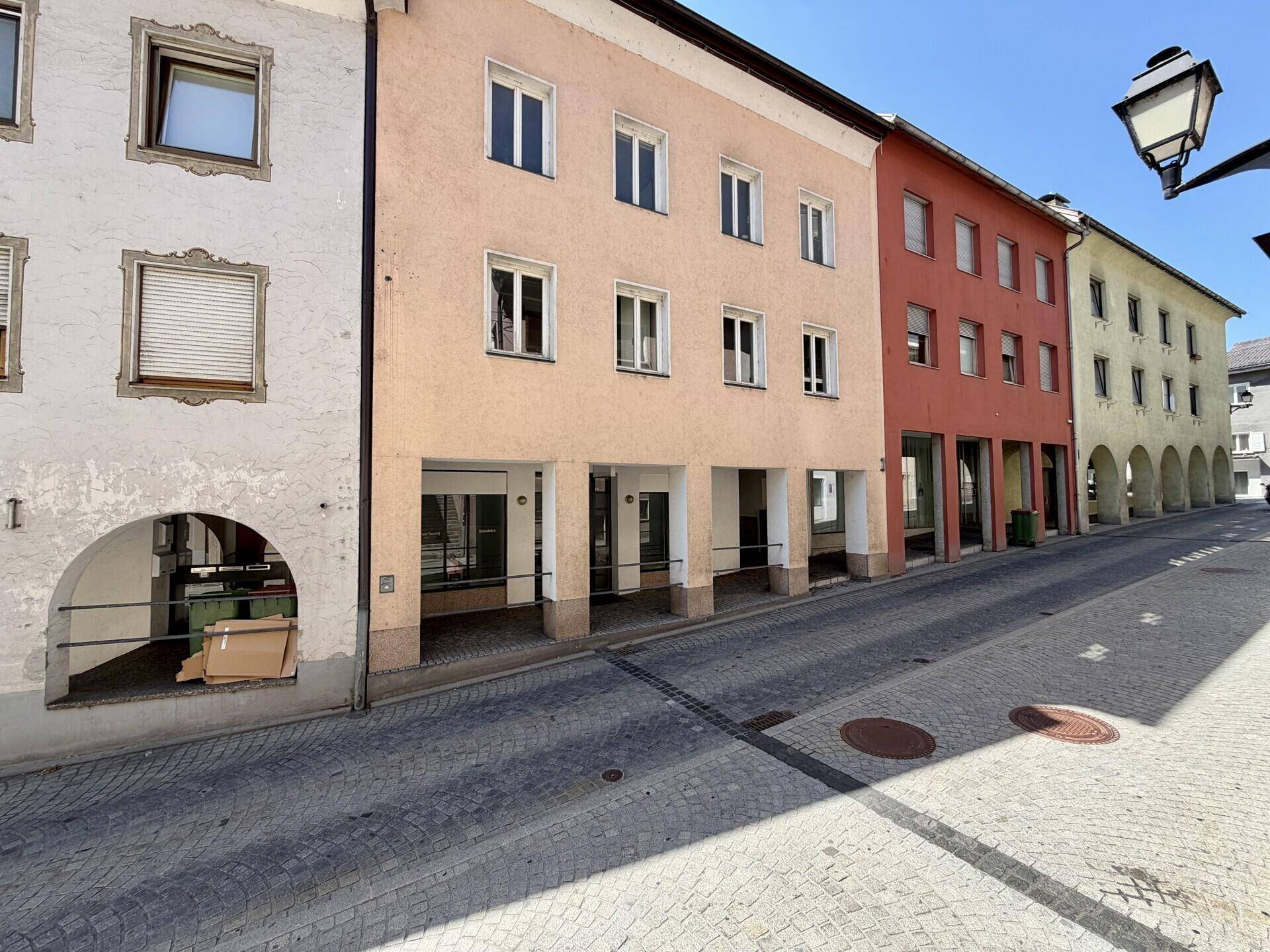 Historische Straße mit mehreren Gebäuden in Reihe, darunter ein hellbeiges Haus mit Arkaden im Erdgeschoss.