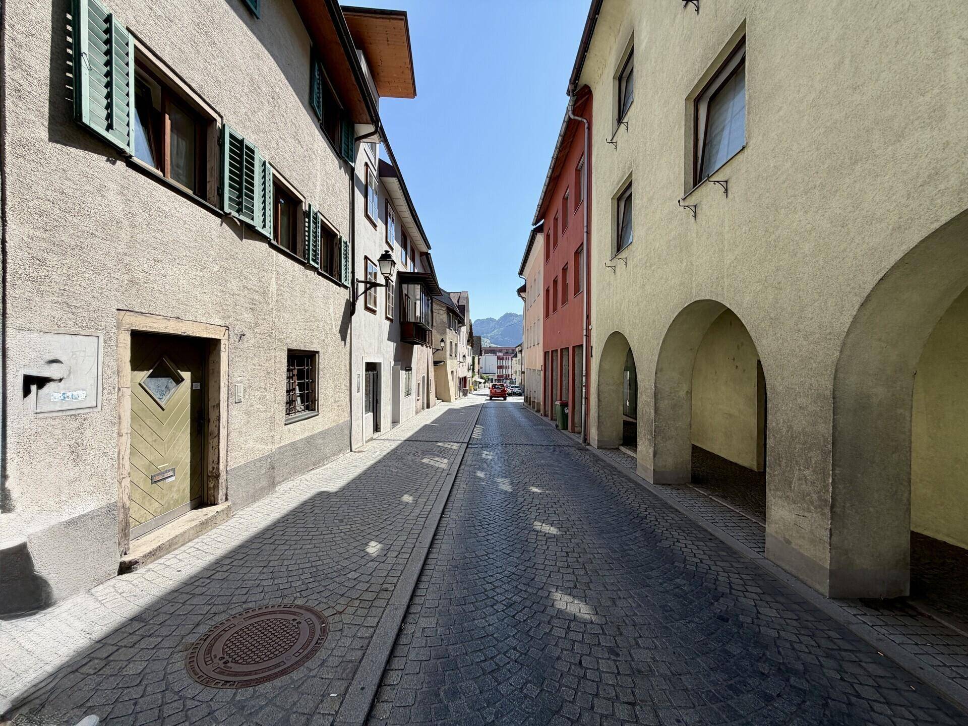 Enge Kopfsteinpflasterstraße in einer historischen Stadt, umgeben von alten Gebäuden und mit Bergblick in der Ferne.