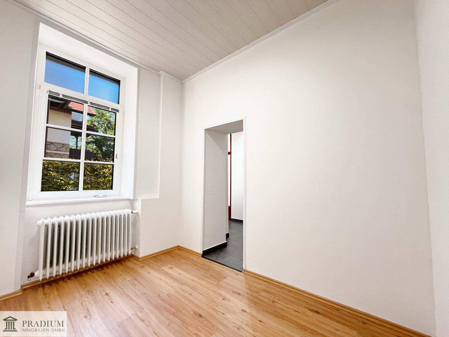 Helles Zimmer mit Laminatboden, großem Fenster und Heizkörper unter dem Fenster.