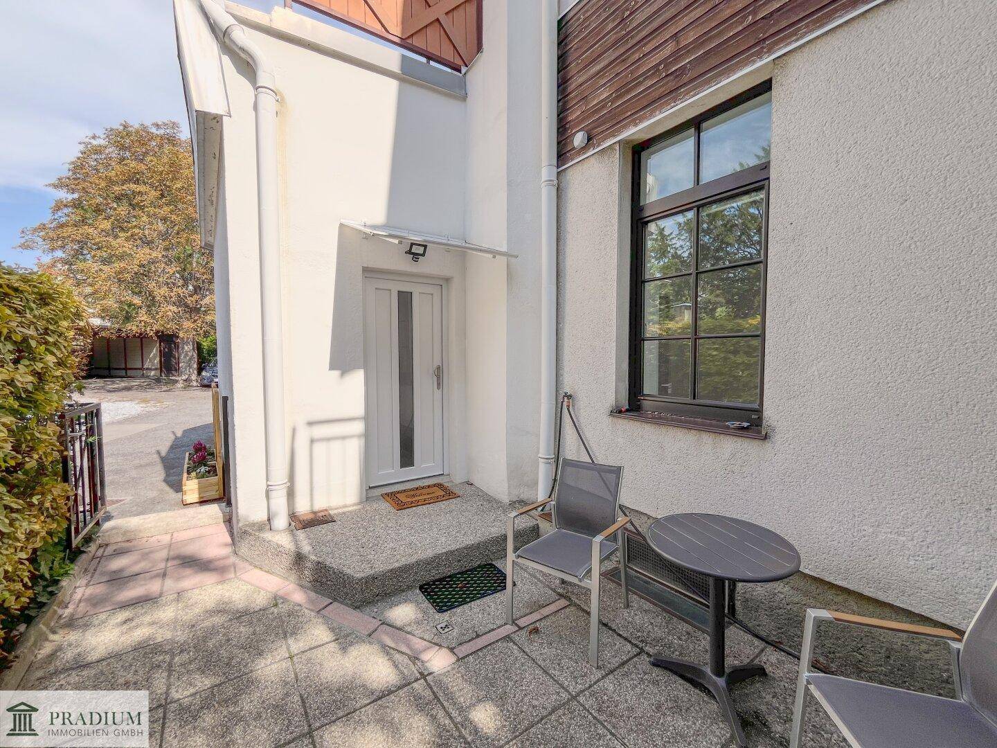 Kleine Terrasse mit Tisch und Stühlen neben dem Hauseingang, umgeben von Grün.