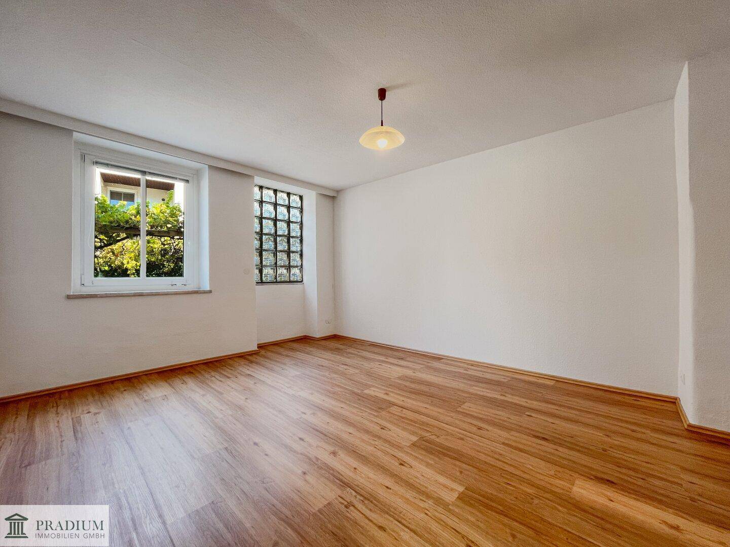 Geräumiges Zimmer mit Laminatboden und zwei Fenstern, eines davon mit Glasbausteinen für Privatsphäre.
