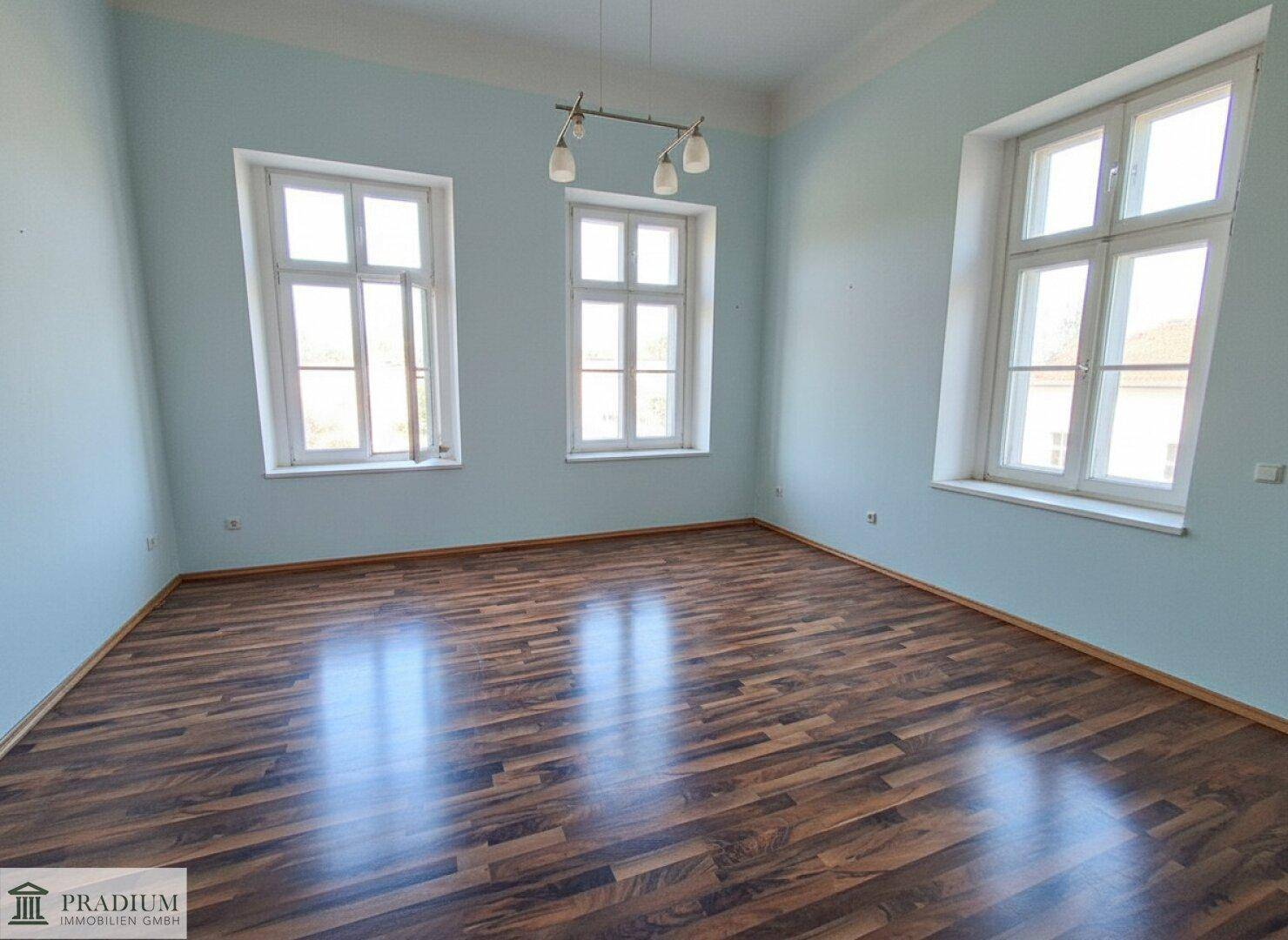 Leerer Raum mit Holzboden, hellblauen Wänden und großen Fenstern.