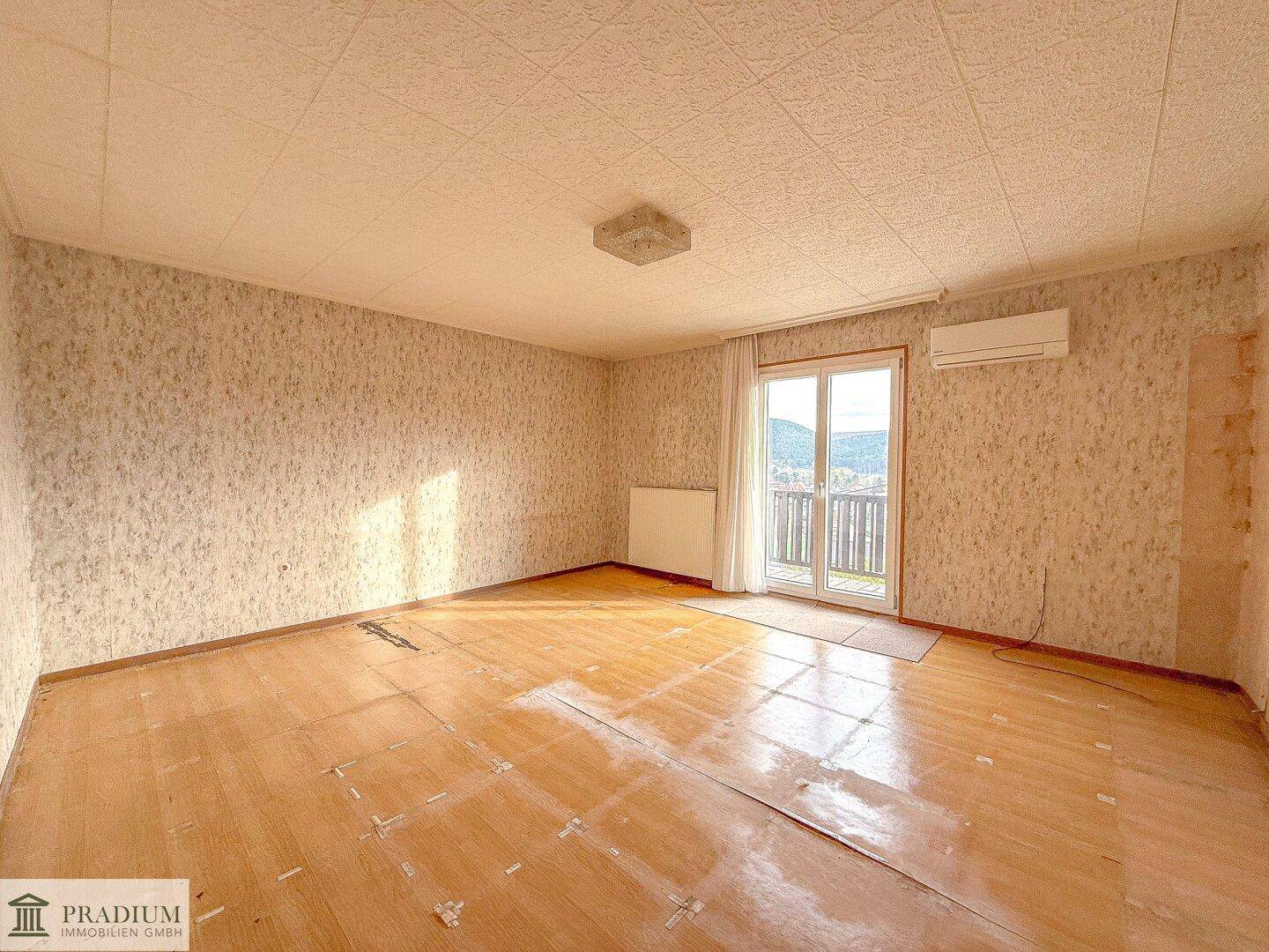 Heller Wohnraum mit Holzboden, Zugang zum Balkon und Blick auf die umliegende Landschaft, renovierungsbedürftig.