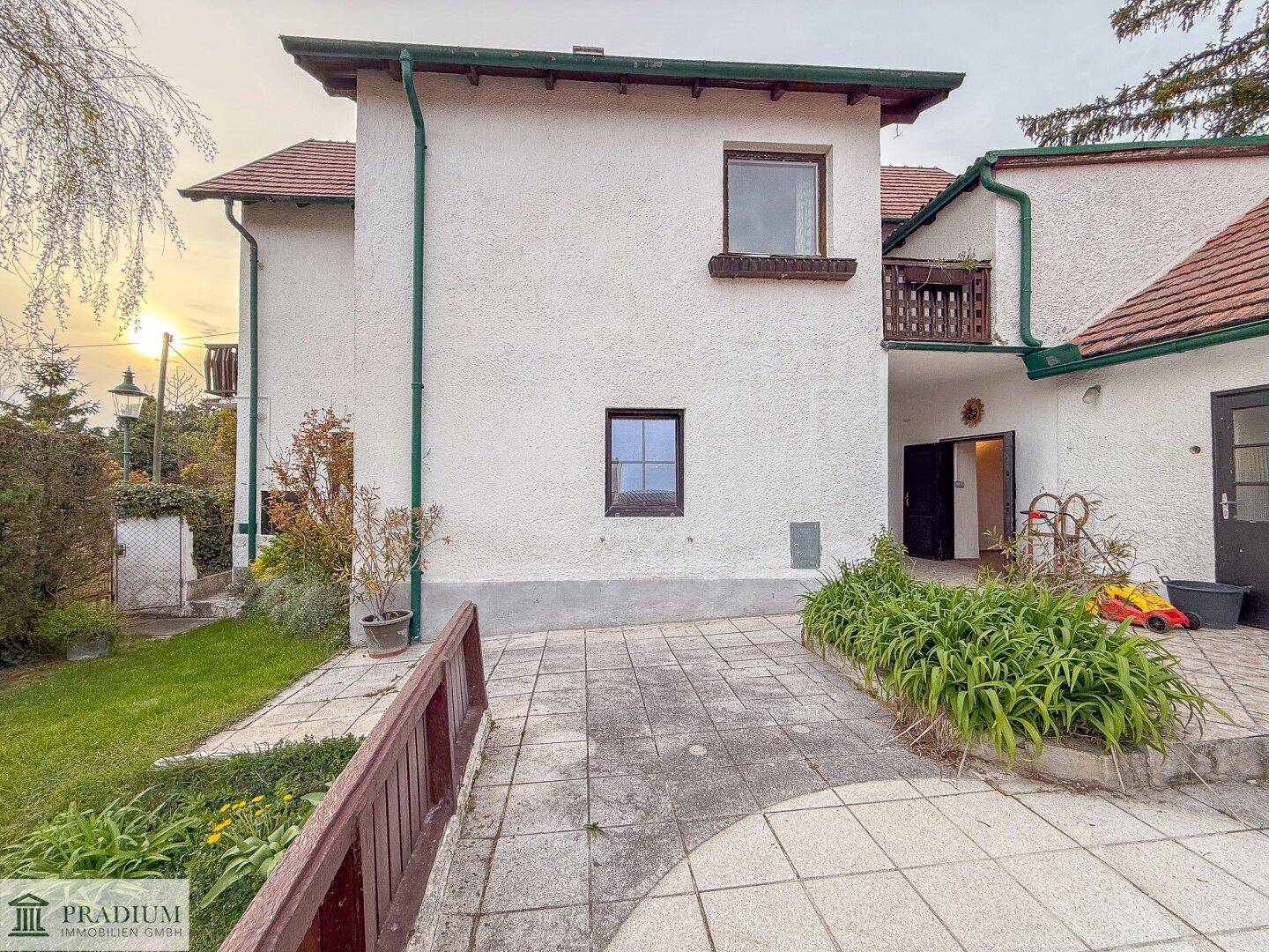 Seitenansicht des Hauses mit weißer Fassade, kleinen Fenstern und gepflastertem Zugangsbereich.