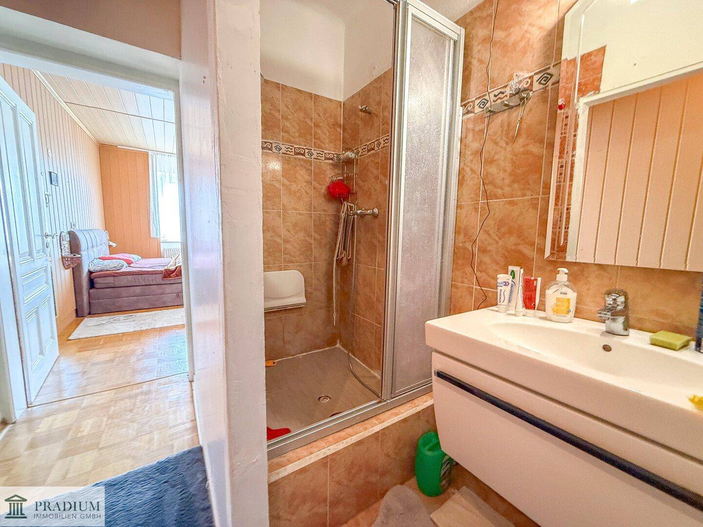 Badezimmer mit braunen Fliesen, einer Duschkabine und einem Waschbecken, angrenzend an ein Schlafzimmer.