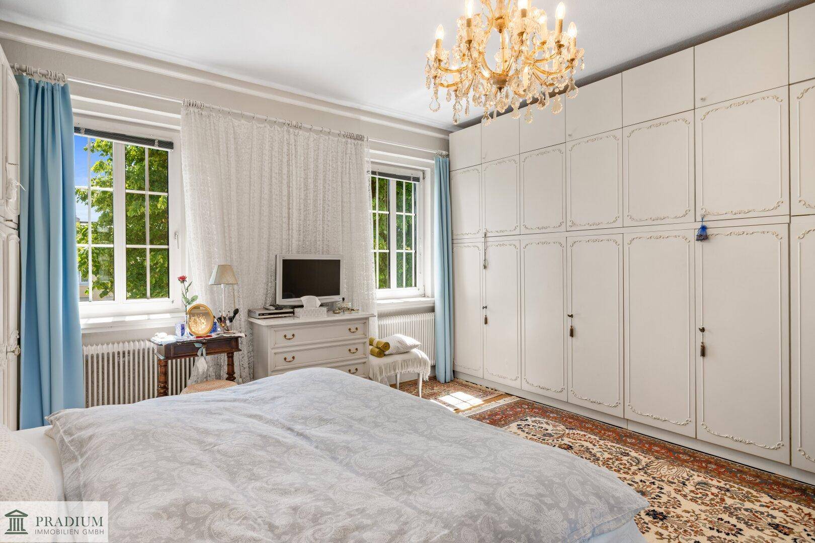 Helles Schlafzimmer mit großen Einbauschränken, klassischem Kronleuchter und Blick ins Grüne.