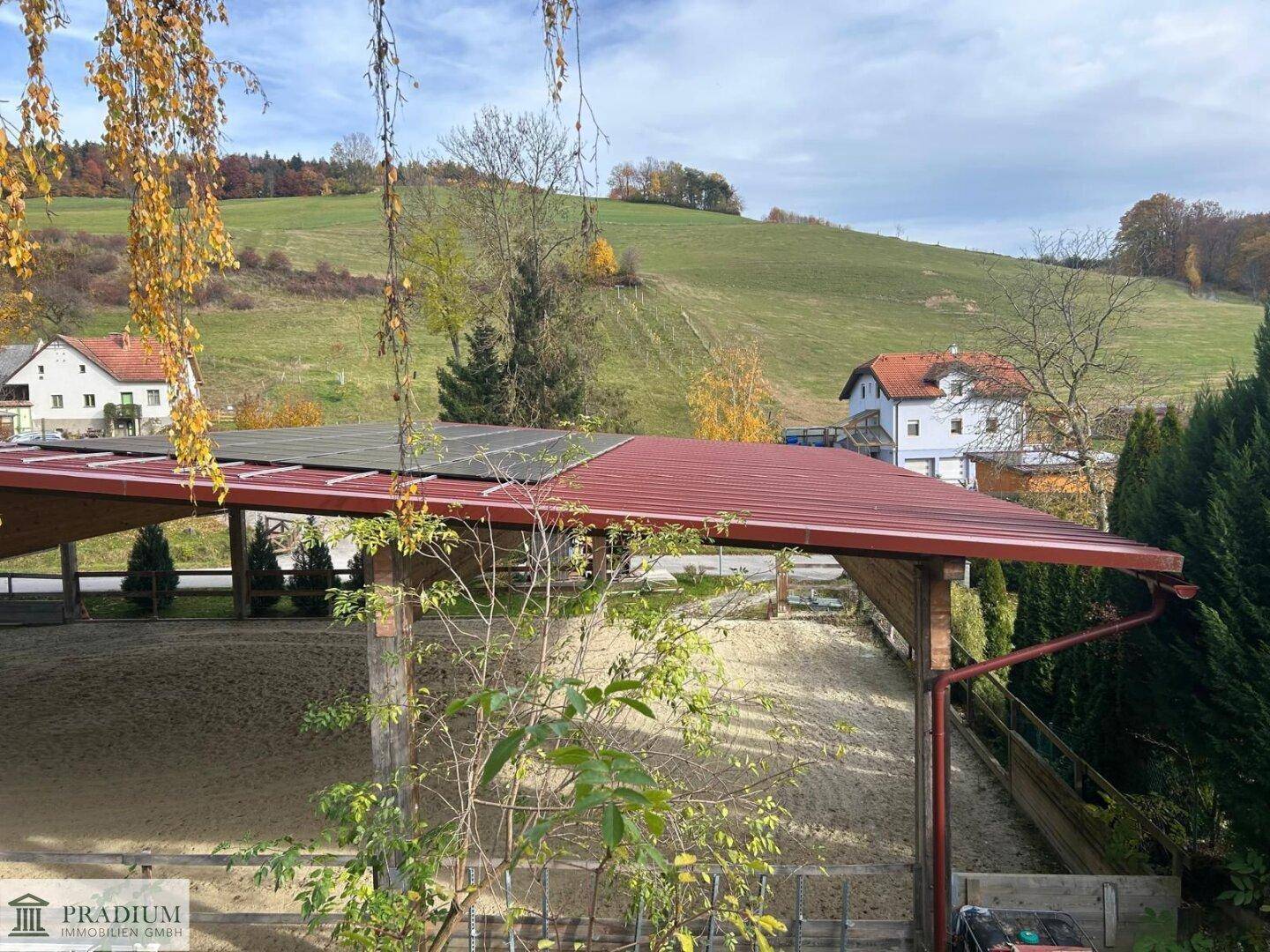 Überdachte Reitanlage mit Solarpaneelen auf dem Dach, eingebettet in eine hügelige Landschaft.