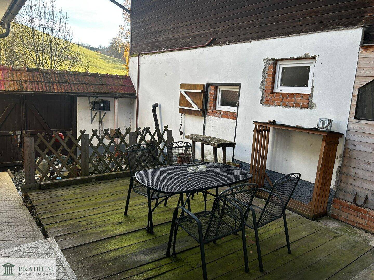 Rustikale Holzterrasse mit Metall-Gartengarnitur neben einem älteren Gebäude.