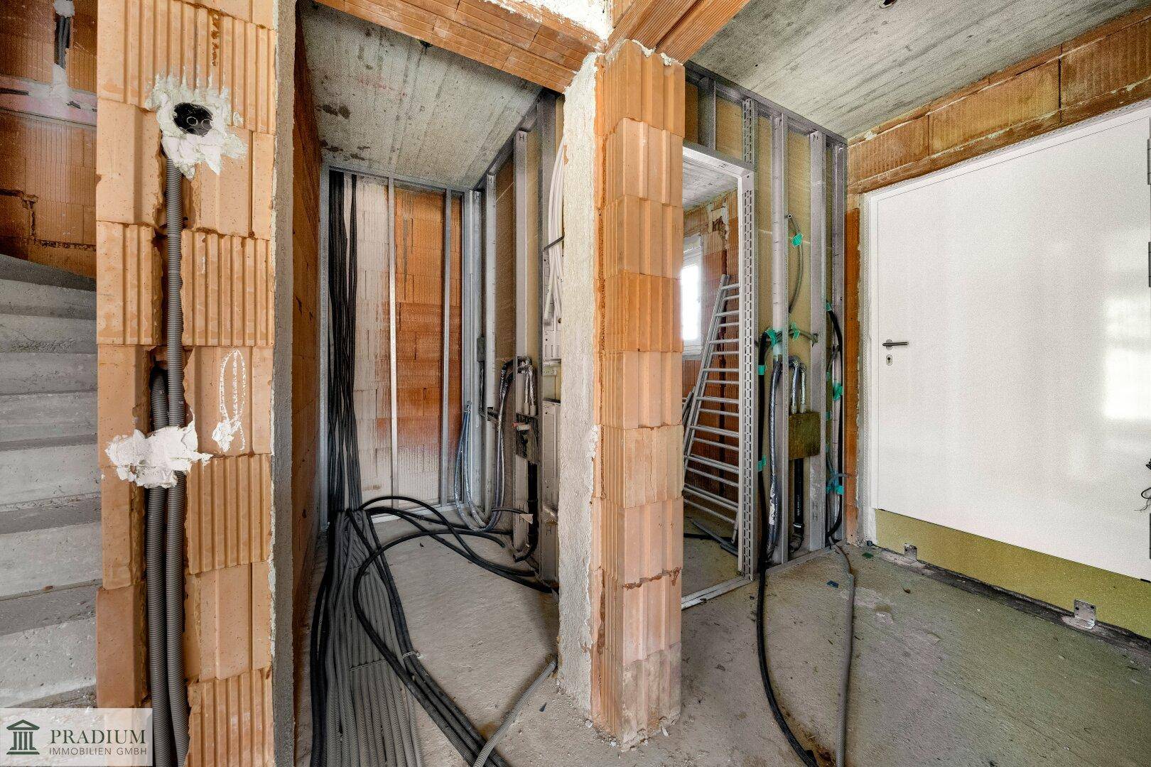 Flurbereich im Rohbau mit Treppenaufgang, sichtbaren Installationen und einer Tür.