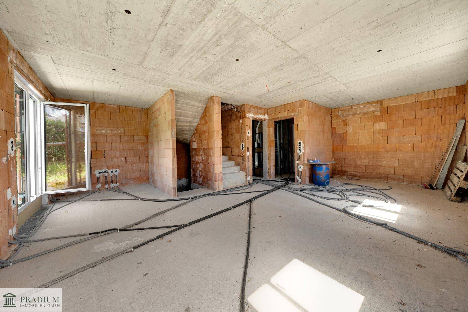 Großer Rohbau-Raum mit Treppenaufgang, Betonboden und sichtbaren Elektroinstallationen.