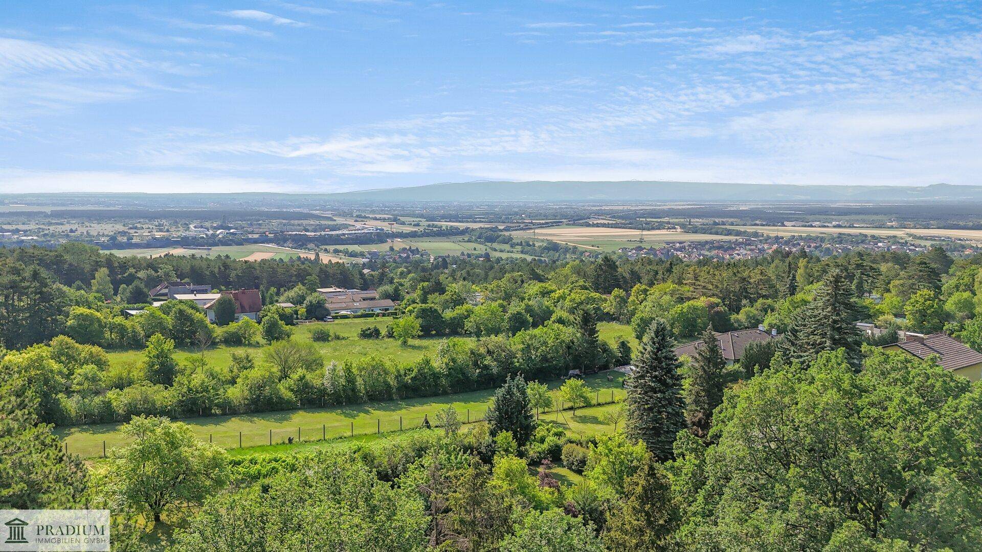 Weitläufiger Panoramablick über die grüne Landschaft und entfernte Siedlungen unter blauem Himmel.