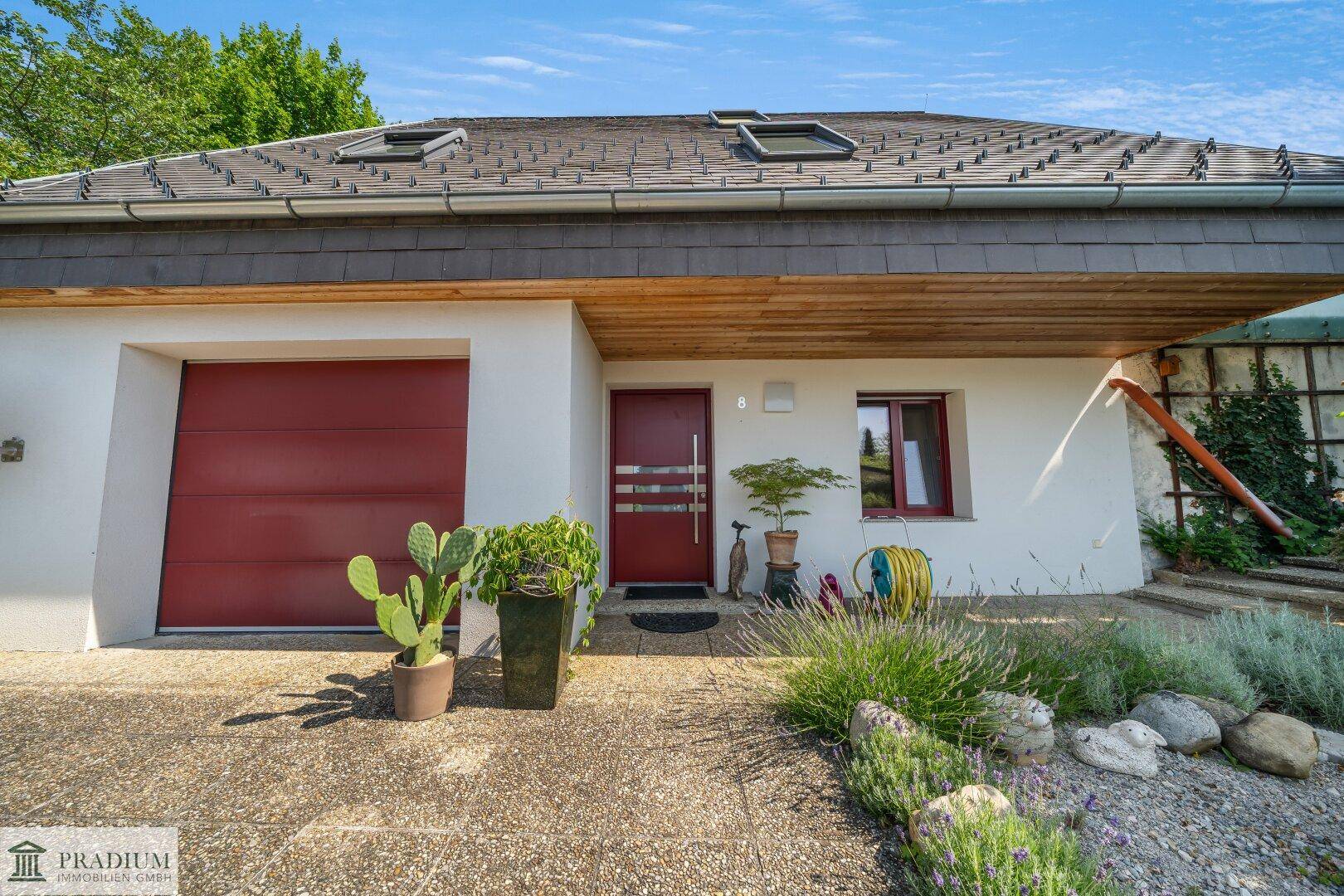 Eingangsbereich des Hauses mit roter Garagentür und Haustür, umgeben von Pflanzen.