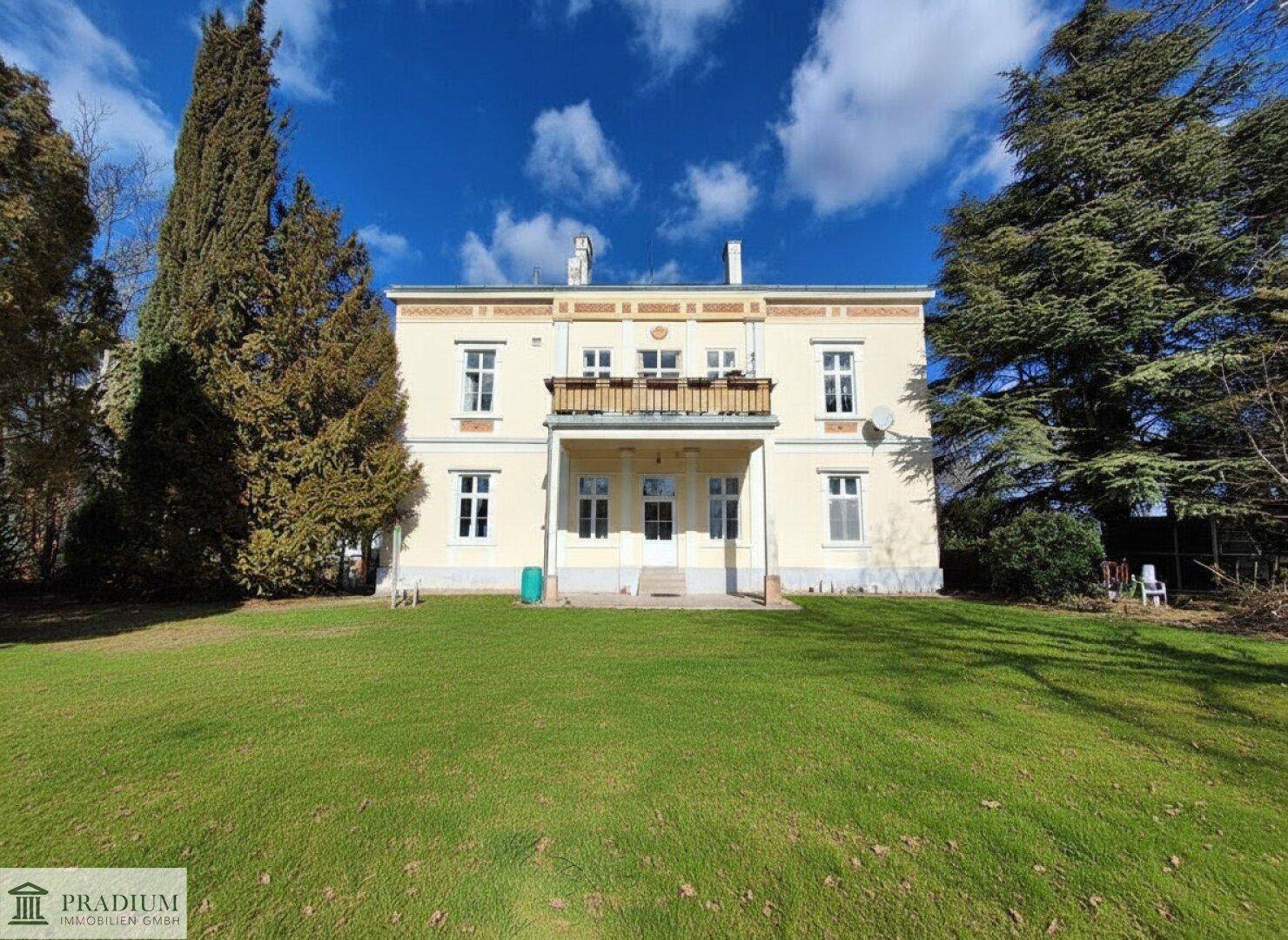 Eindrucksvolle gelbe Villa mit klassischer Architektur, umgeben von einem weitläufigen Rasen und hohen Bäumen.