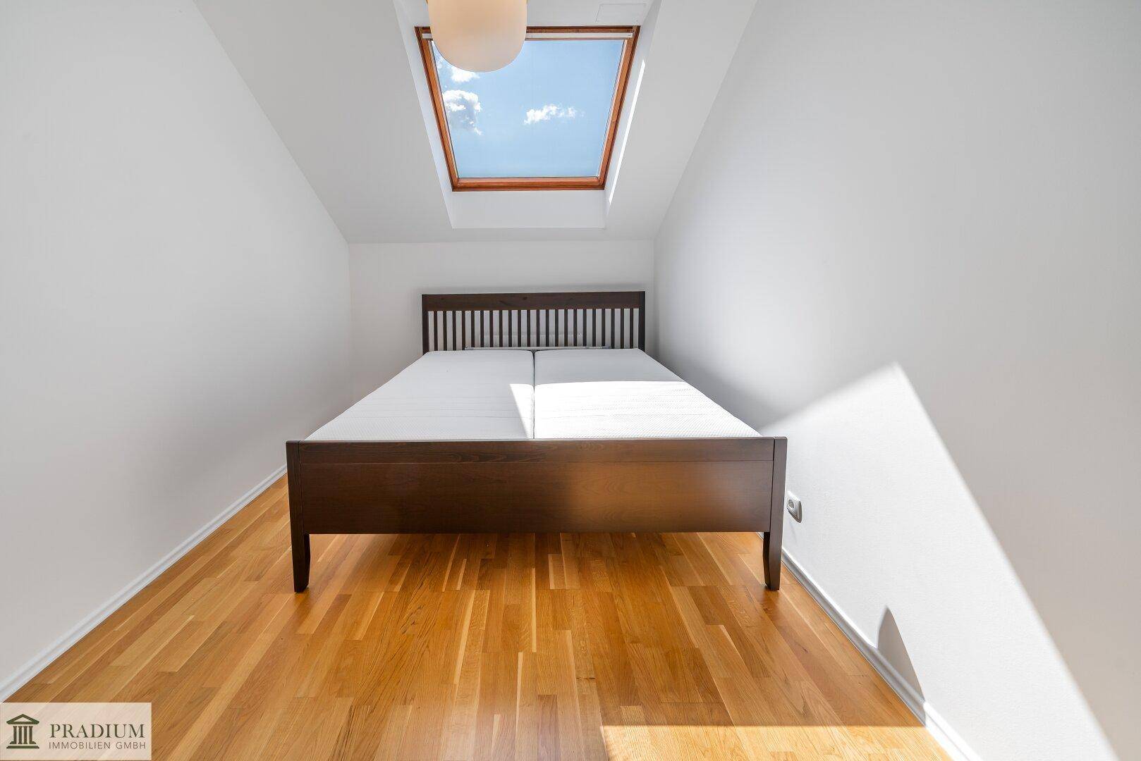 Helles Schlafzimmer im Dachgeschoss mit Holzbett und großem Dachfenster für natürliche Belichtung.