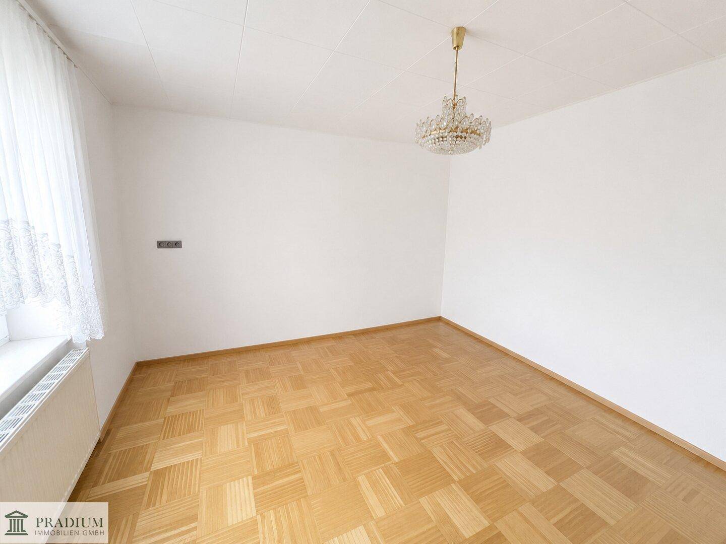 Leerer Raum mit klassischem Parkettboden, weißen Wänden und einem Kronleuchter an der Decke.