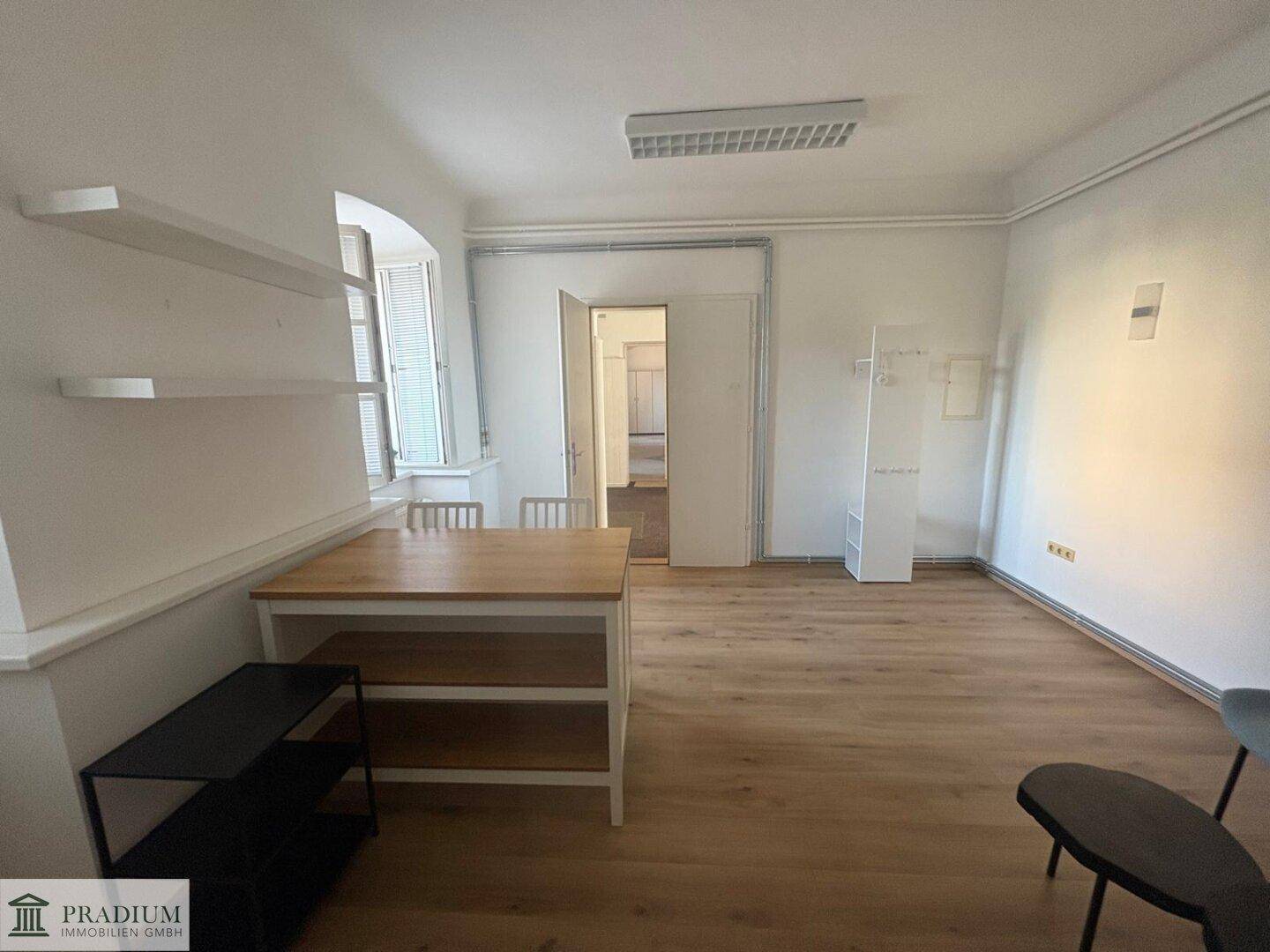 Heller Büroraum mit Holzboden, Regalen und einem Blick in den angrenzenden Flur.