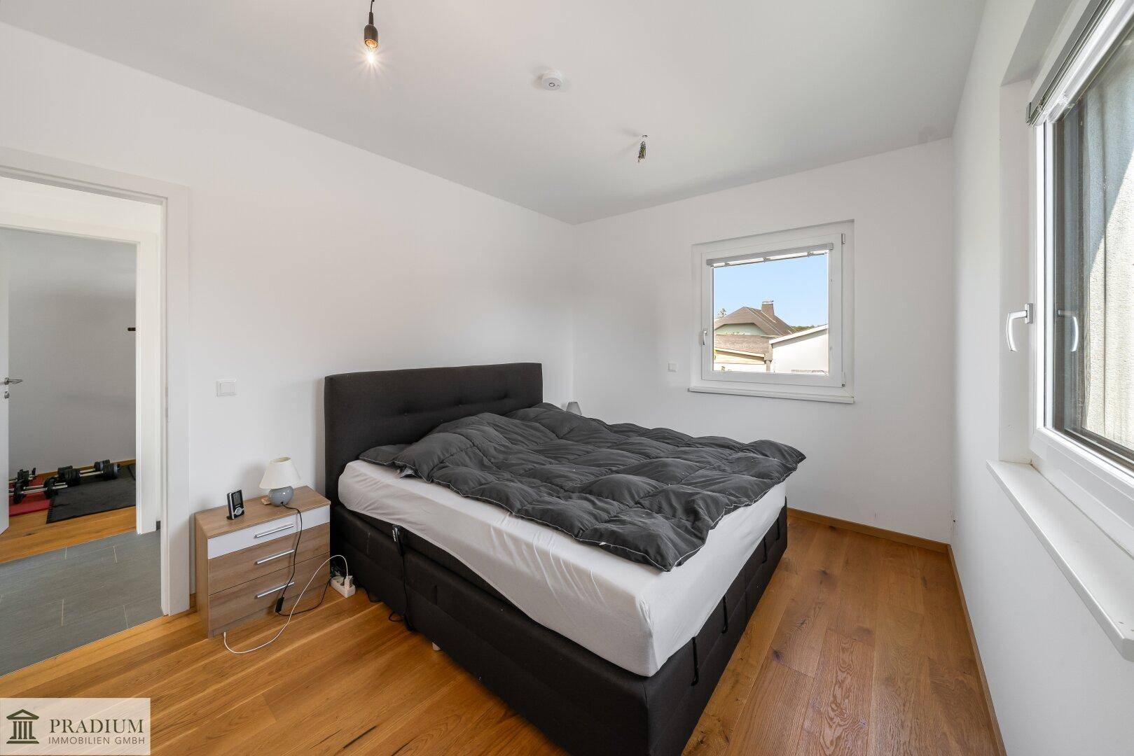 Schlafzimmer mit dunklem Bett, Holzboden und Fenster, das viel Tageslicht hereinlässt.