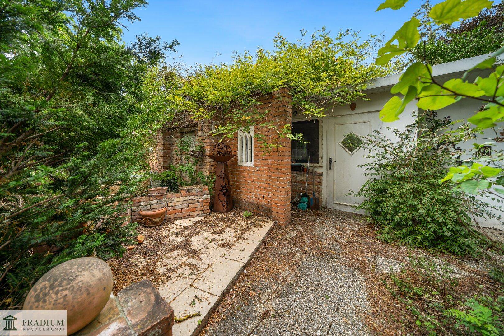Kleines Gartenhaus aus Ziegeln, umrankt von Pflanzen, mit einem gepflasterten Zugang und weißer Tür.