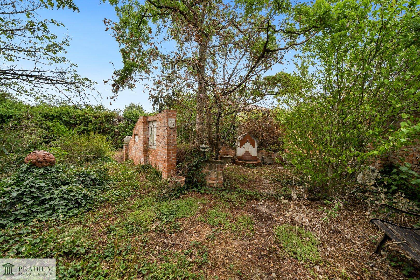 Verwilderter Gartenbereich mit alten Ziegelstrukturen, dichtem Grün und einer Gartenbank.