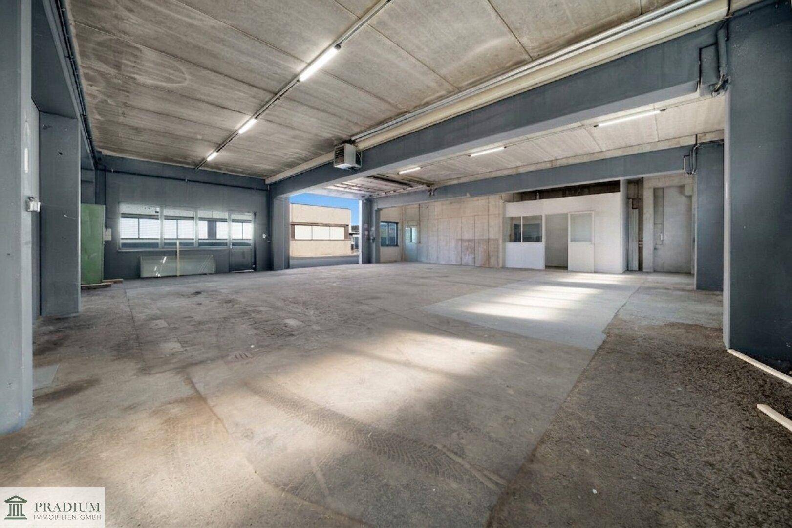 Große Lagerhalle mit Betonboden, hohen Decken und Tageslicht durch Fenster.