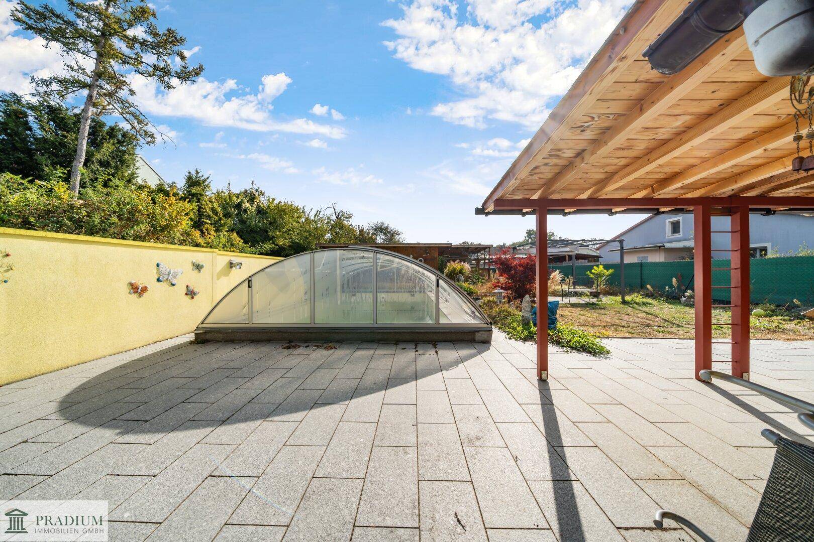 Große gepflasterte Terrasse mit Überdachung und einem überdachten Pool im Hintergrund unter blauem Himmel.
