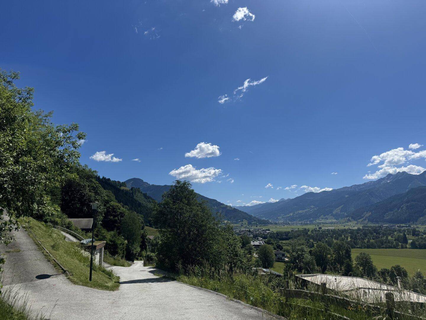 Blick von einem gewundenen Weg in ein grünes Tal mit Bäumen und fernen Bergen unter blauem Himmel.