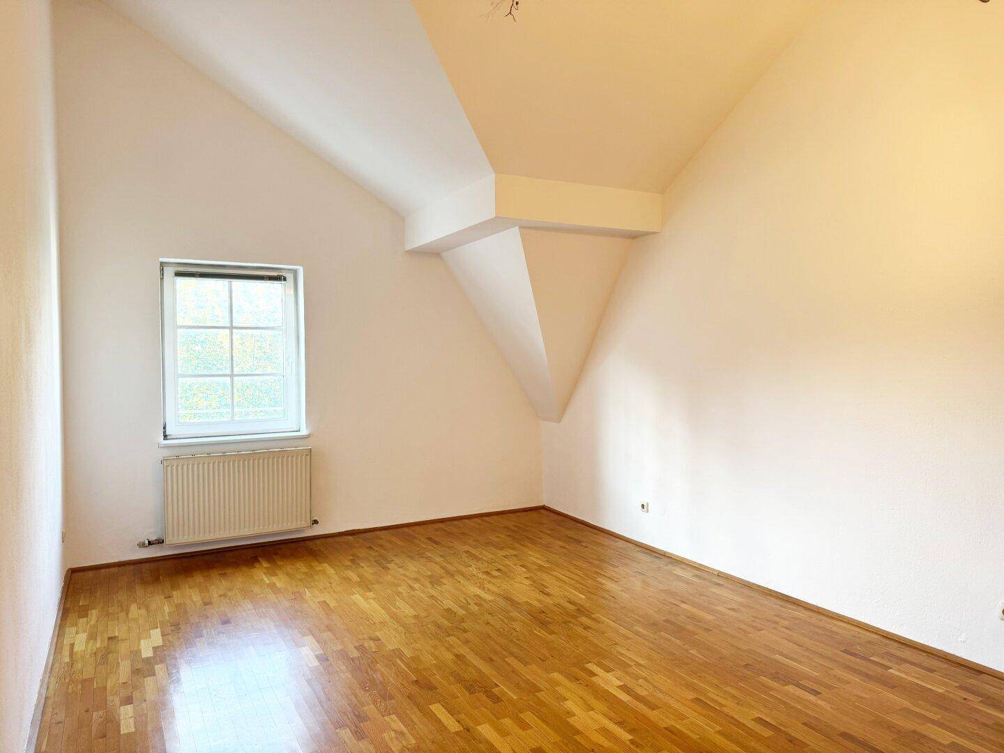 Geräumiger, heller Raum mit Dachschräge, Parkettboden und einem Fenster.