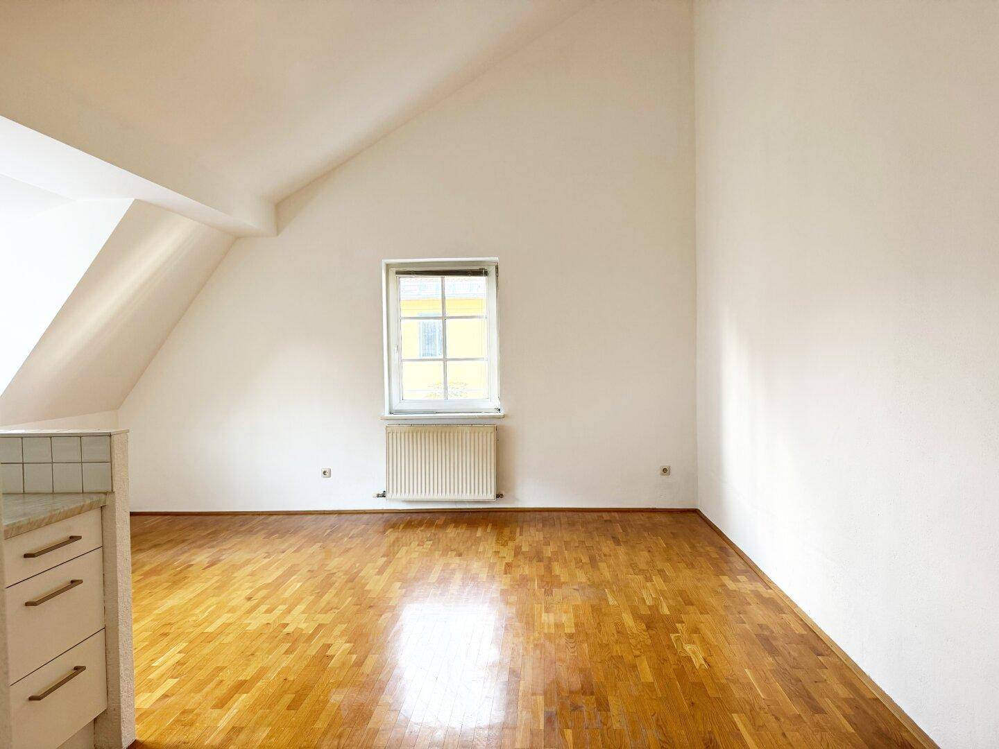 Heller, leerer Raum mit Dachschräge, Parkettboden und einem Fenster.