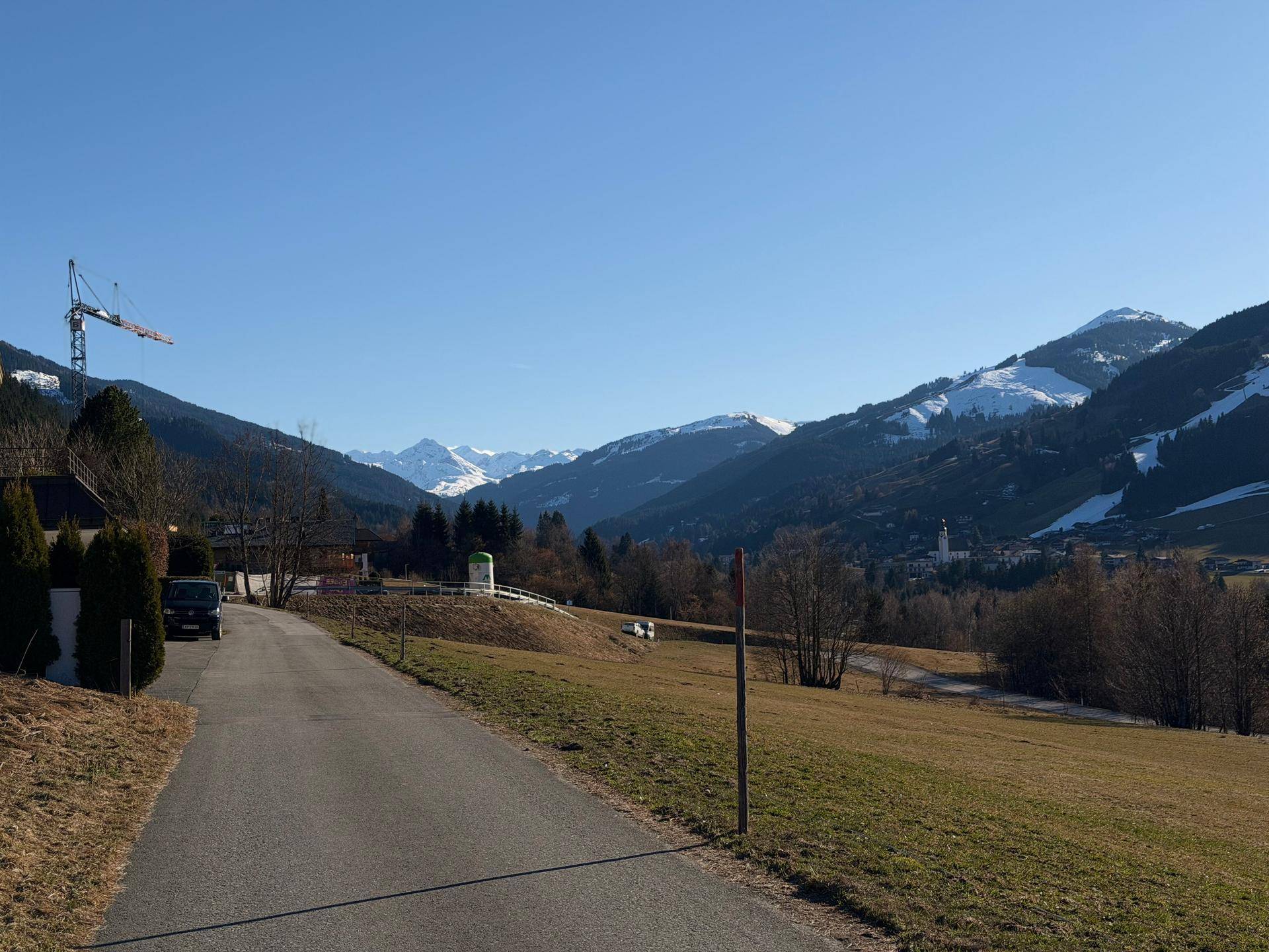 Ländliche Straße führt durch grüne Hügel mit schneebedeckten Bergen und einem Kran im Hintergrund.